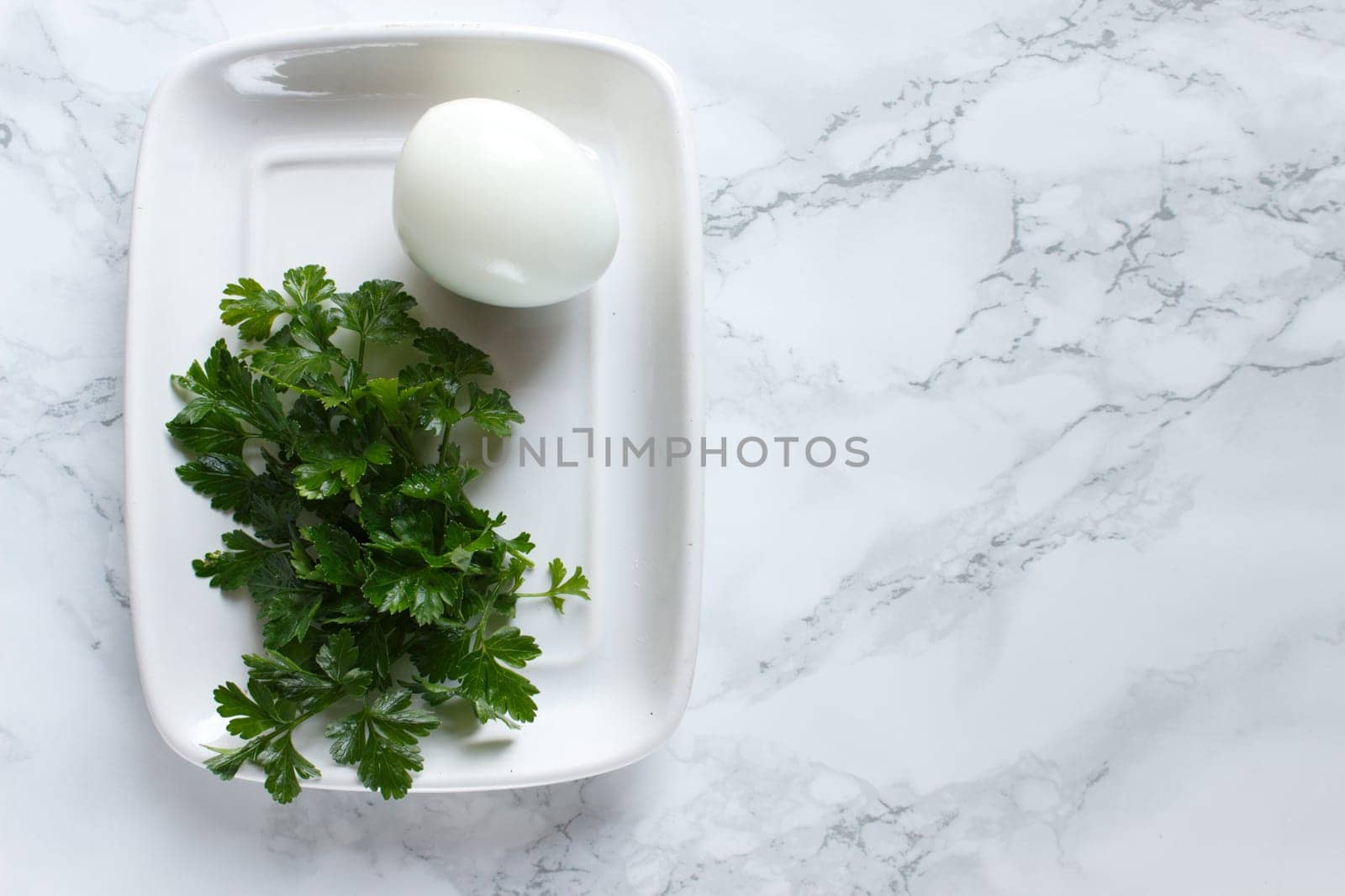 Boiled chicken egg and parsley on a white plate on a marble background.