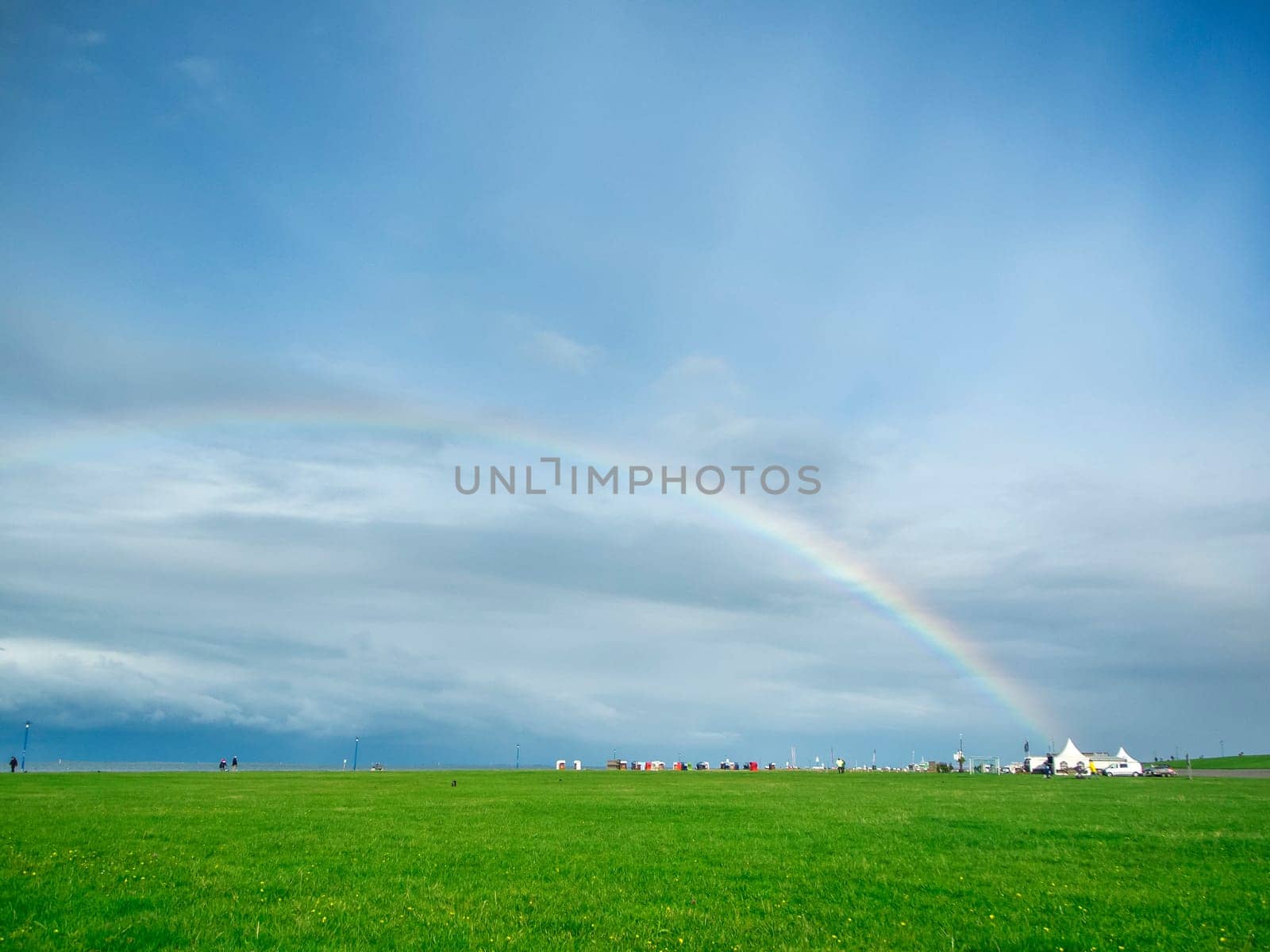 Landscape view on the Lower Saxony coast with snack tents next to a green hang gliding meadow under an overcast sky with a rainbow.