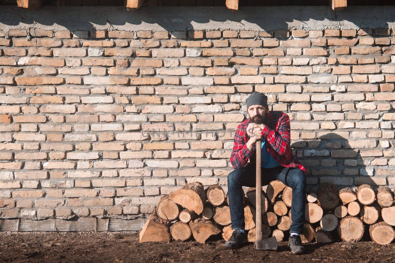 The sad and tired lumberjack worker with axe is sitting on firewood on brick wall.