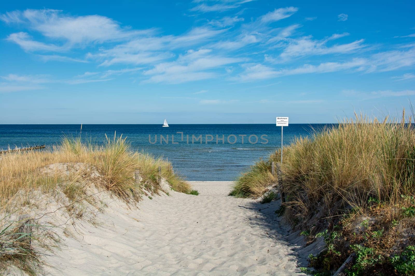 Path between the sand dunes overlooking the sea and a sailing ship and blue sky. With sign "Grones - Please keep your distance - risk of accident"""