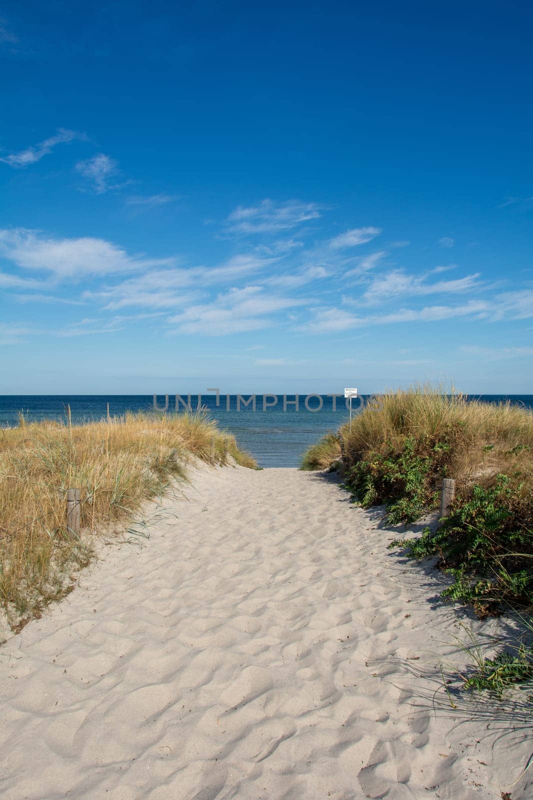 Path between the sand dunes overlooking the sea with blue sky. With sign "Grones - Please keep your distance - risk of accident"""