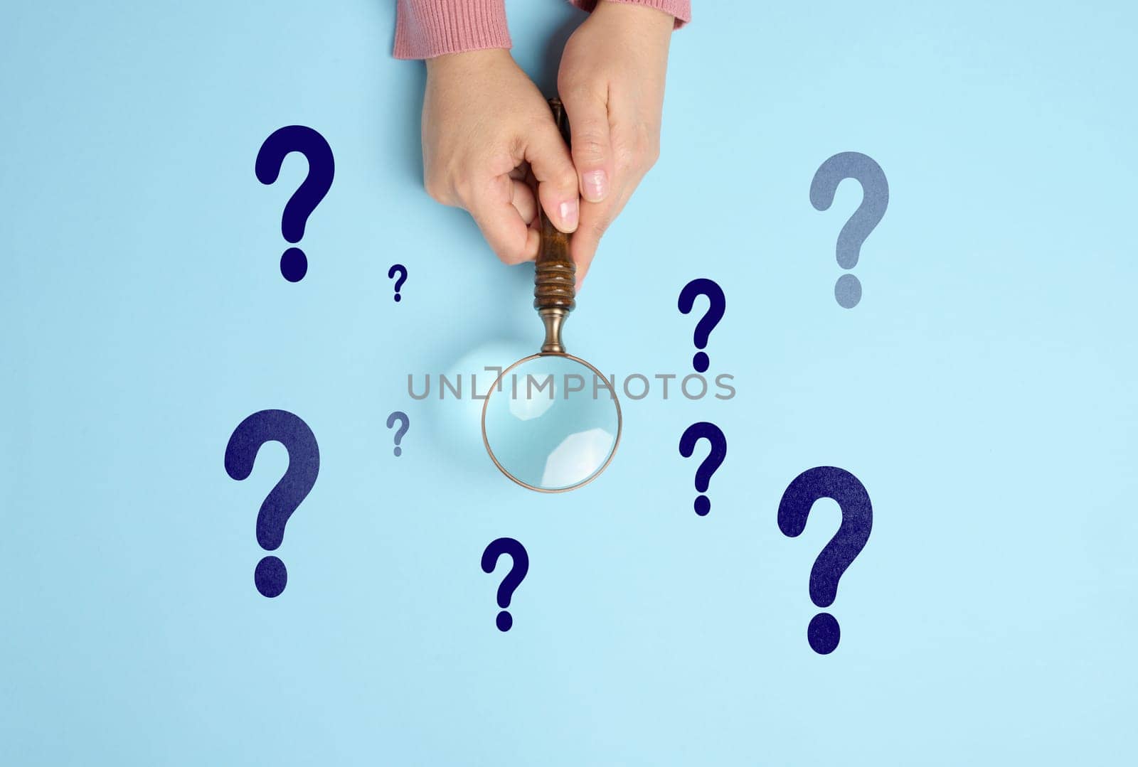 Female hand holds a wooden magnifying glass and question marks on a blue background. The concept of finding an answer to questions, truth and uncertainty.