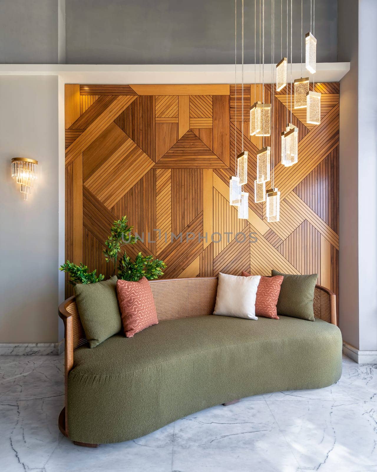 A stylish modern green couch sits in a modern living room with a warm decorated wood cladding wall and a chandelier hanging from the ceiling. The room is decorated with plants and tasteful accessories