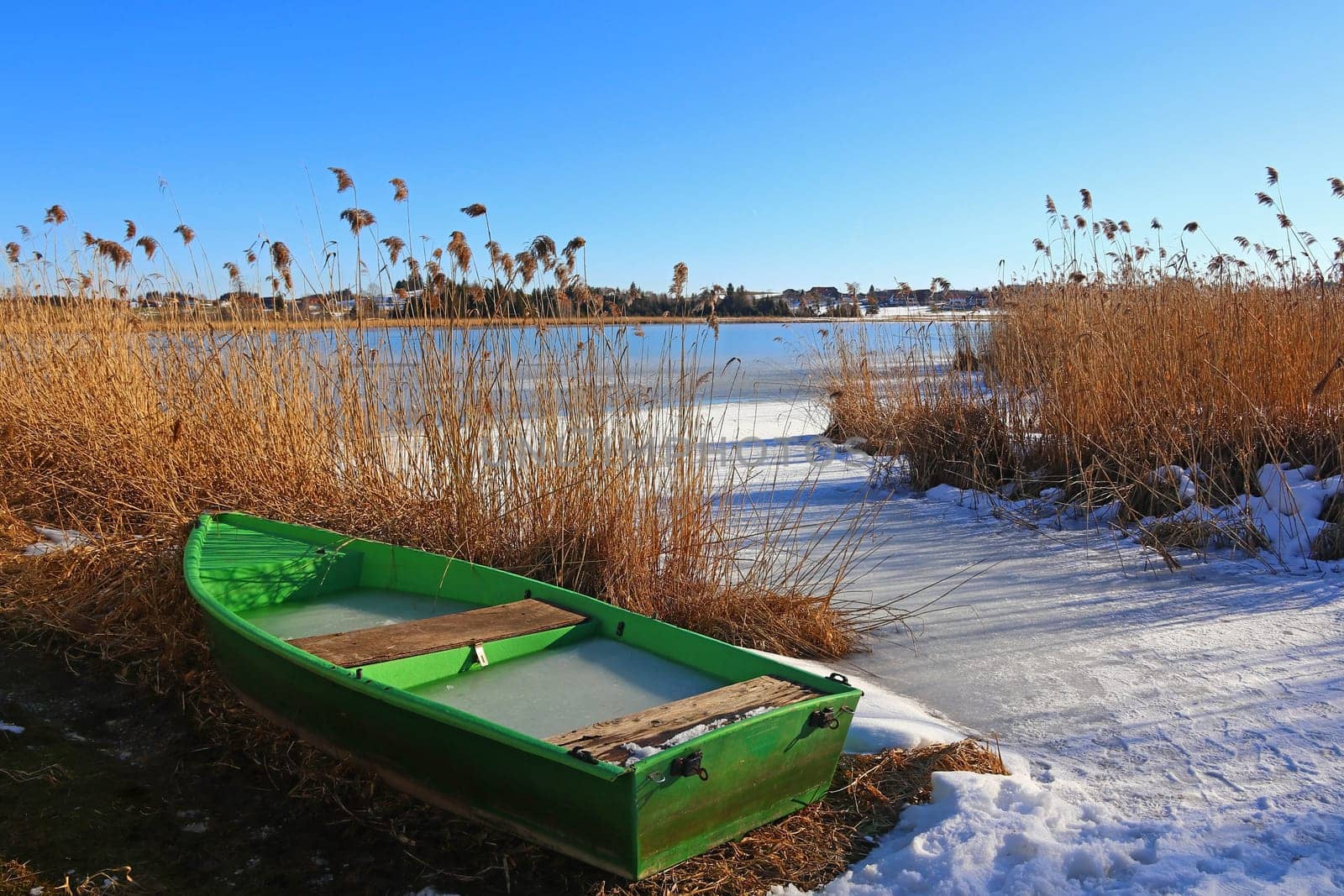 A green boat lies on the shore of a frozen lake in winter
