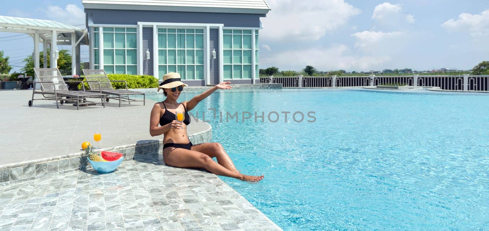 A woman is enjoying a sunny day sitting on the edge of a swimming pool, holding a glass of refreshing fruit juice.