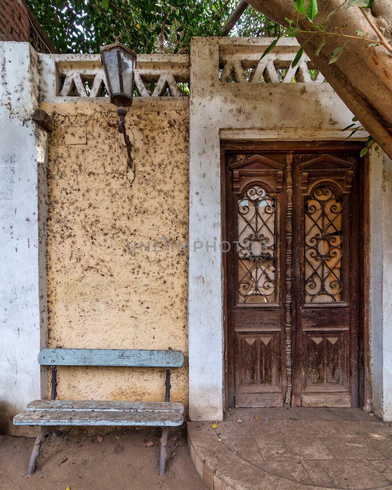 Weathered wooden blue bench sits invitingly beside of a classic wooden decorated grunge door. A vintage metal lamp hangs above the bench, adding to the nostalgic charm of the scene