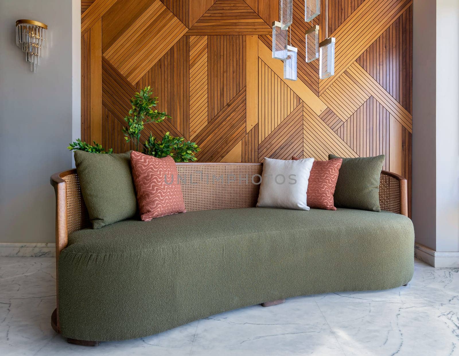 Stylish modern green couch sits in a contemporary living room with decorated wood cladding wall and a chandelier hanging from the ceiling. The room is decorated with plants and tasteful accessories