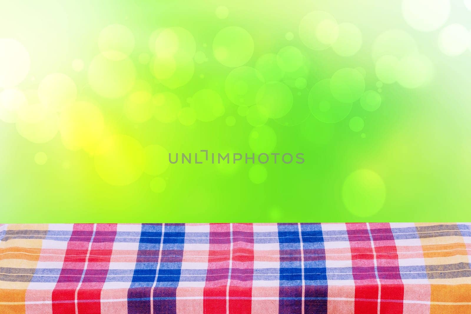 Empty table product. Empty colorful checkered tablecloth over abstract blurred natural green background texture with space for food and product display montage.