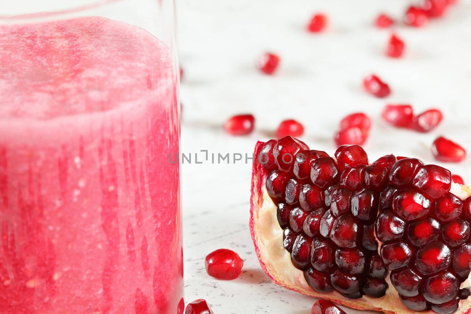 Portion of pomegranate fruit, gem like berries scattered on white board, glass with pink smoothie next to it