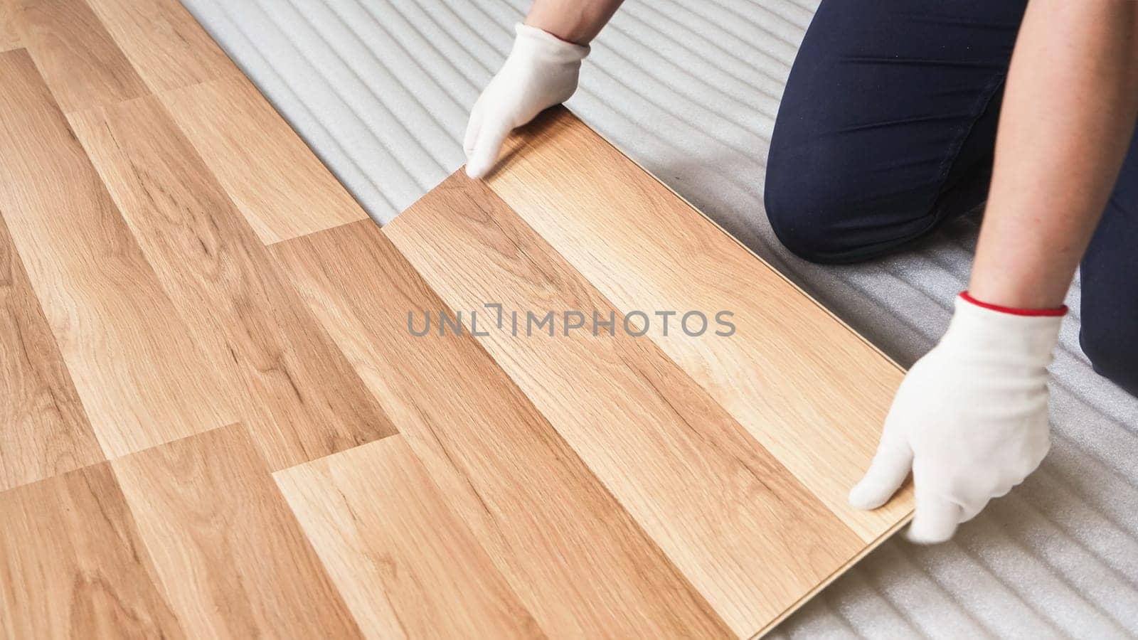 Installing laminated floor, detail on worker hands in white gloves fitting wooden tile, over white foam base layer