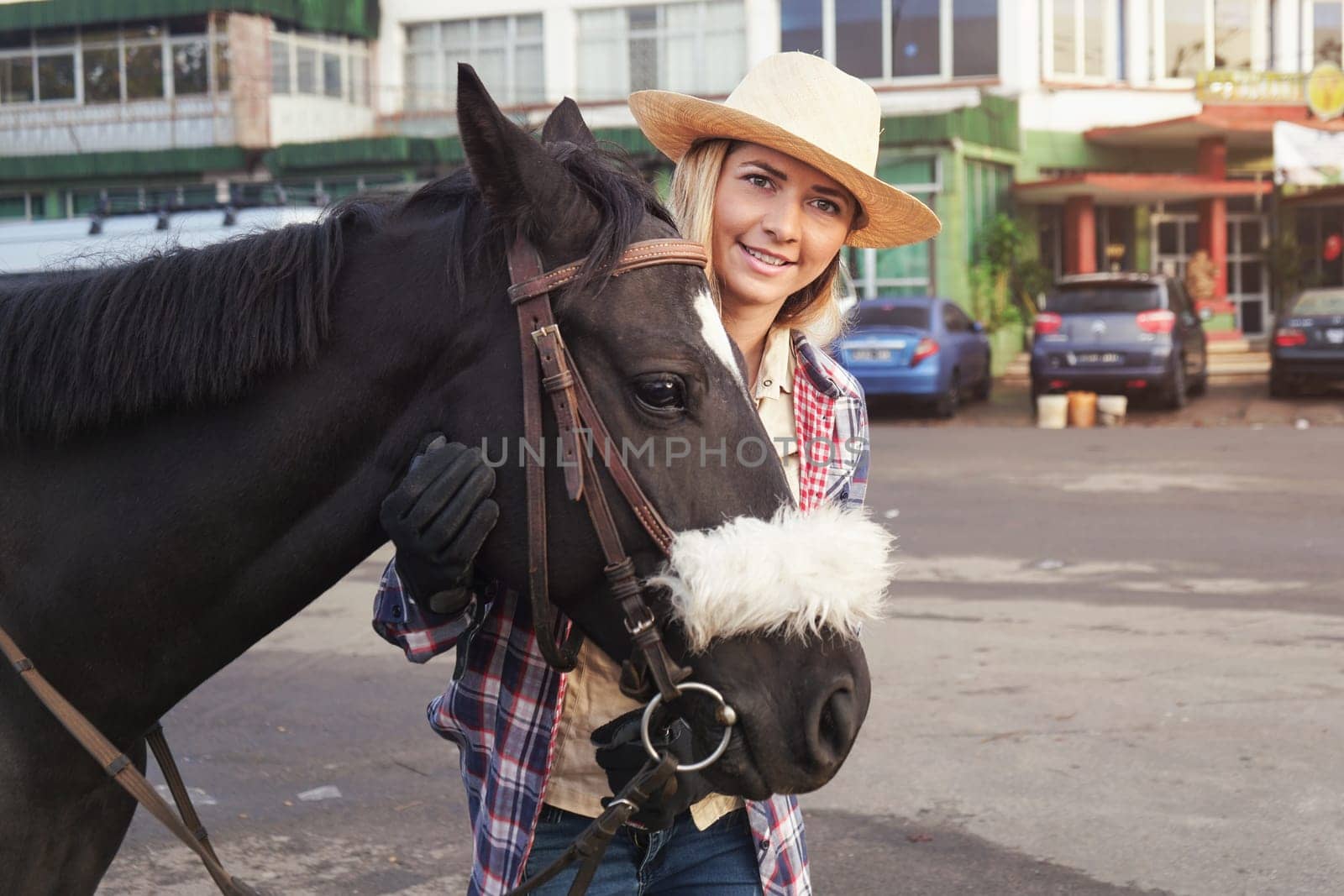 Young woman wearing shirt, gloves and straw hat standing behind dark brown horse, smiling, blurred city street with parked cars in background