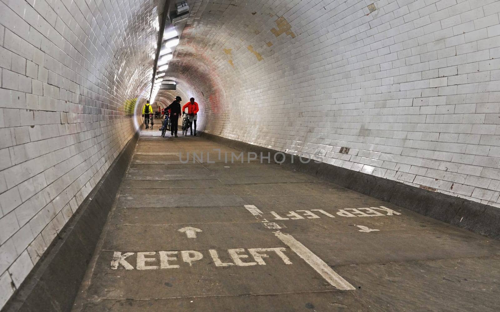 London, United Kingdom - February 02, 2019: Cyclists in bright jackets walking with their bicycles in Greenwich foot tunnel under river Thames, "Keep Left"" sign on the floor"