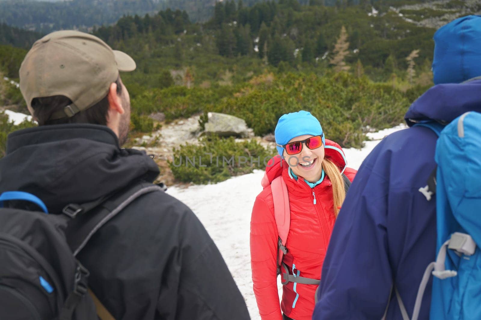 Three hikers wearing jackets, mountains and forest partially covered with snow, young woman in red jacket, blue cap and sunglasses looking to other two, smiling happily, blurred trees at background