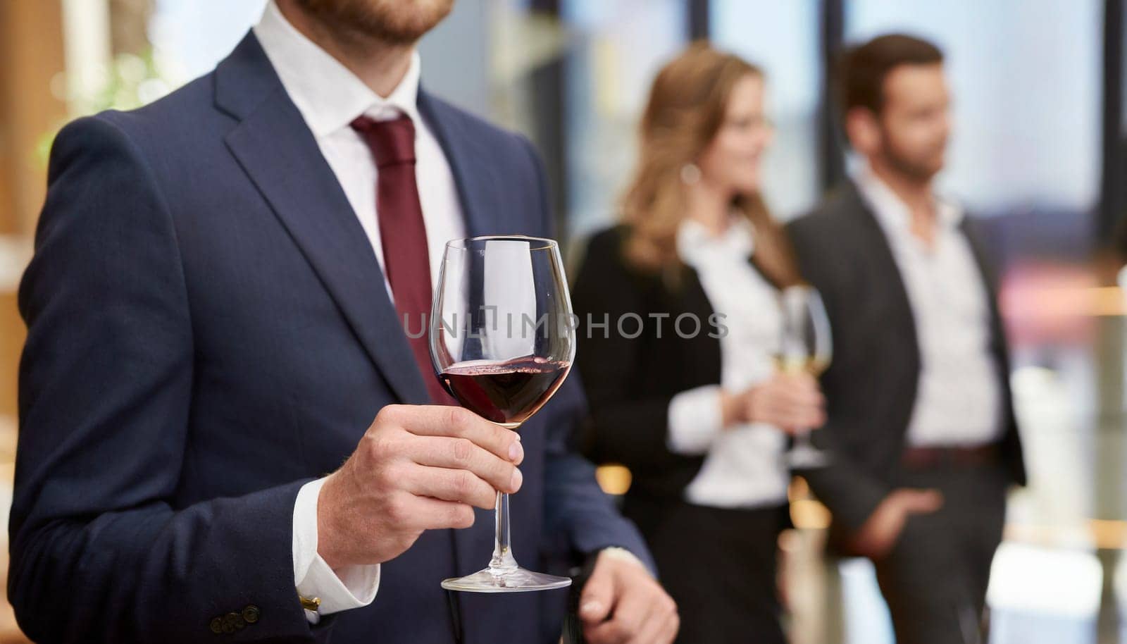 Businessman holding a glass of red wine in the office with colleagues in the background. AI generated