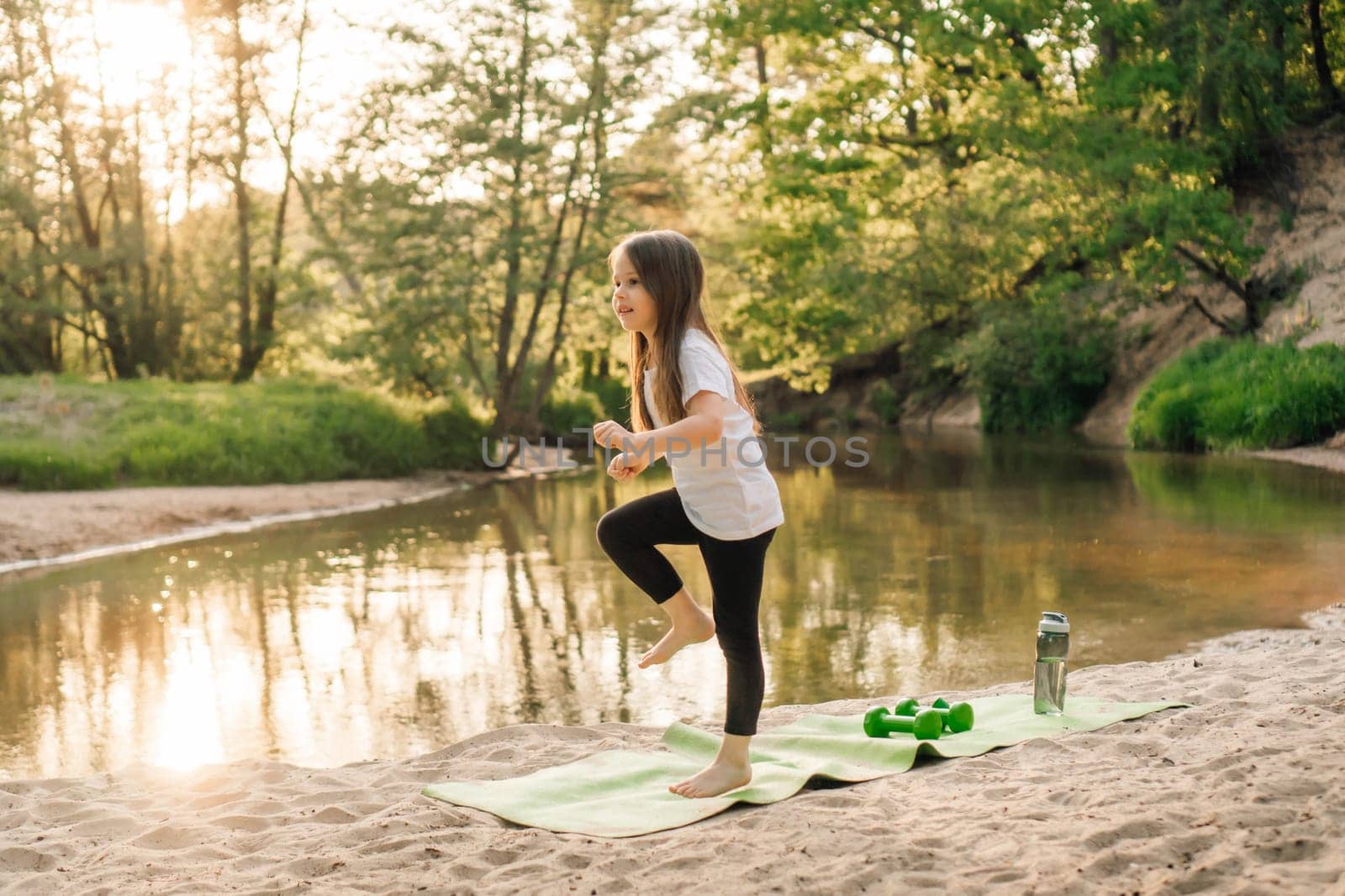 Adorable little female athlete in white t-shirt and black pants standing on green mat near river and doing sports. Energetic girl with brown long hair enjoying activity on sandy beach close to water.