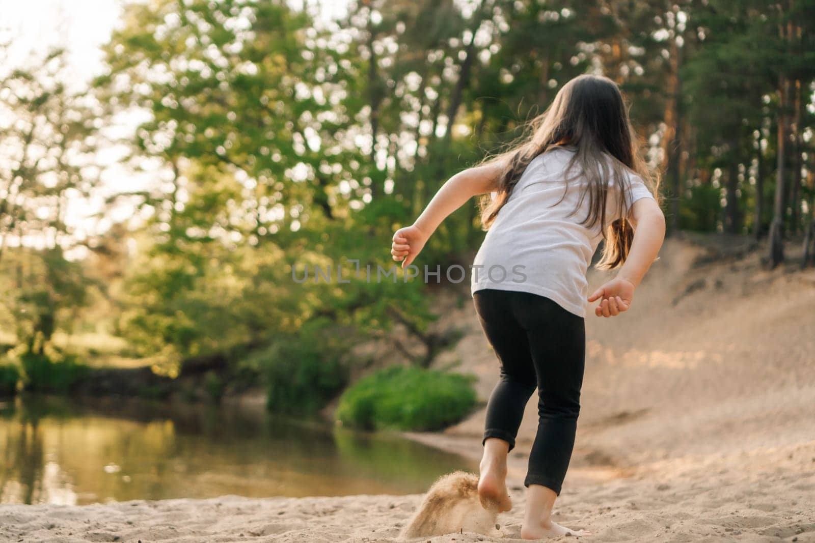 Rear view of little brunette with long hair running on sandy beach near water. Active girl wearing white t-shirt and black leggings jogging in nature close to river.