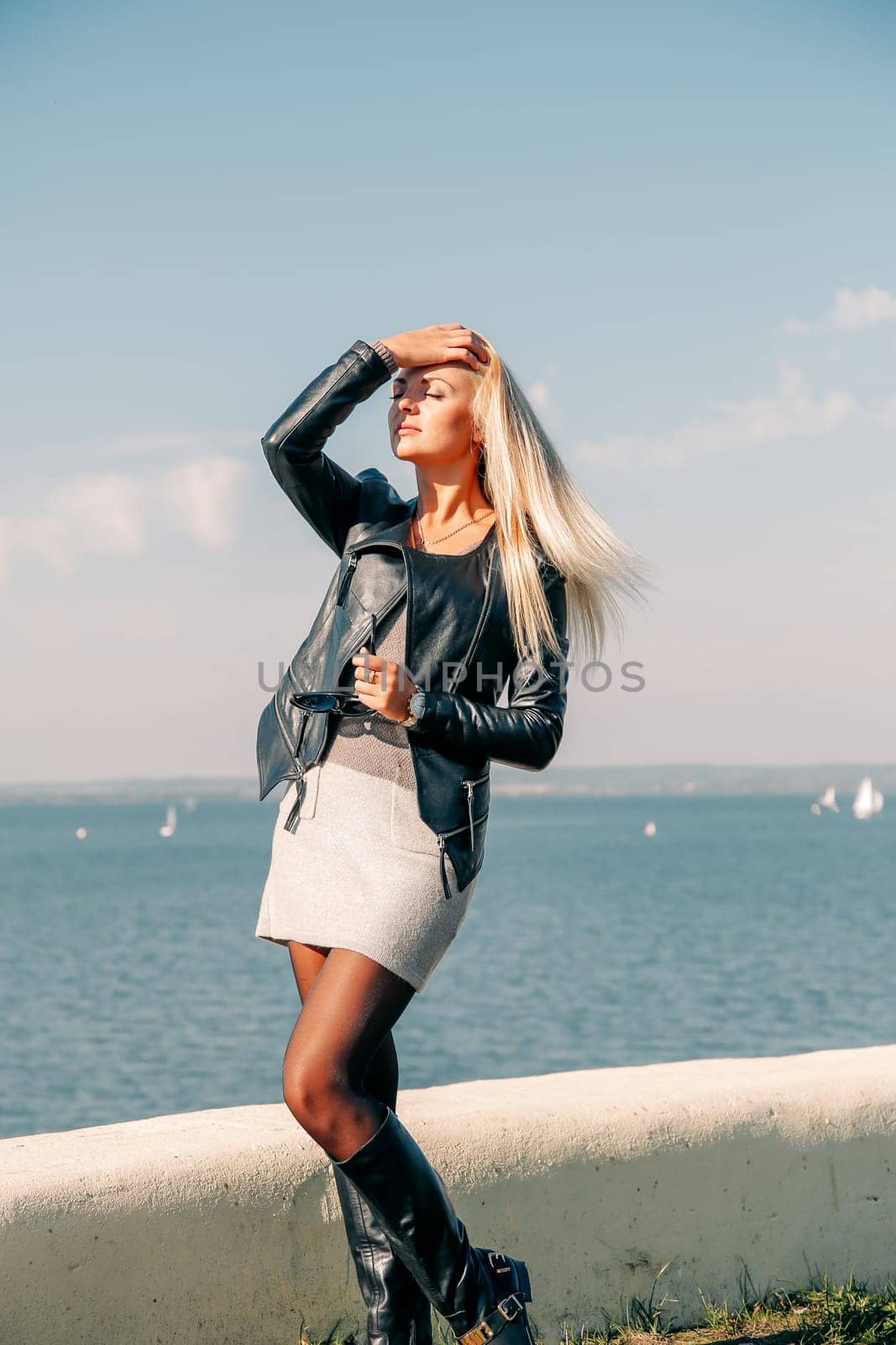 A blonde girl with long hair and glasses poses on the shore of the blue sea in the afternoon