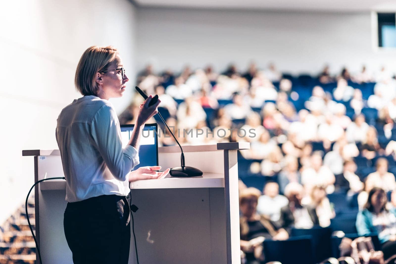 Female speaker giving a talk on corporate business conference. Unrecognizable people in audience at conference hall. Business and Entrepreneurship event