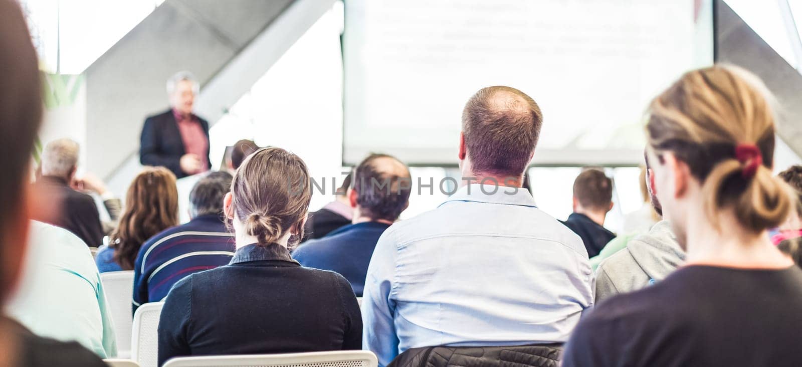 Man giving presentation in lecture hall. Male speeker having talk at public event. Participants listening to lecture. Rear view, focus on people in audience.