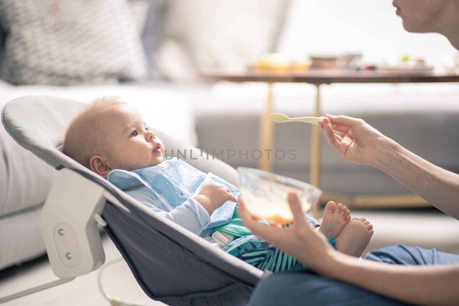 Mother spoon feeding her baby boy child in baby chair with fruit puree. Baby solid food introduction concept