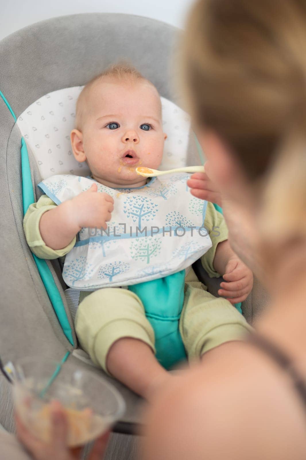 Mother spoon feeding her baby boy child in baby chair with fruit puree. Baby solid food introduction concept