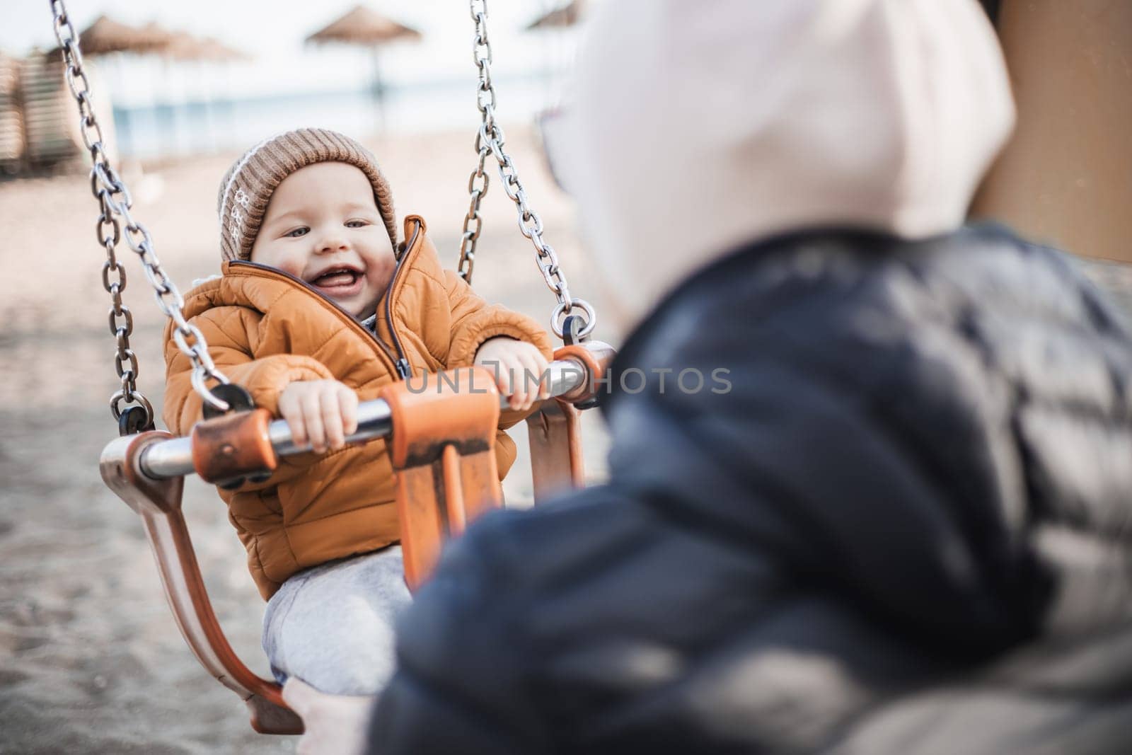 Mother pushing her cheerful infant baby boy child on a swing on sandy beach playground outdoors on nice sunny cold winter day in Malaga, Spain