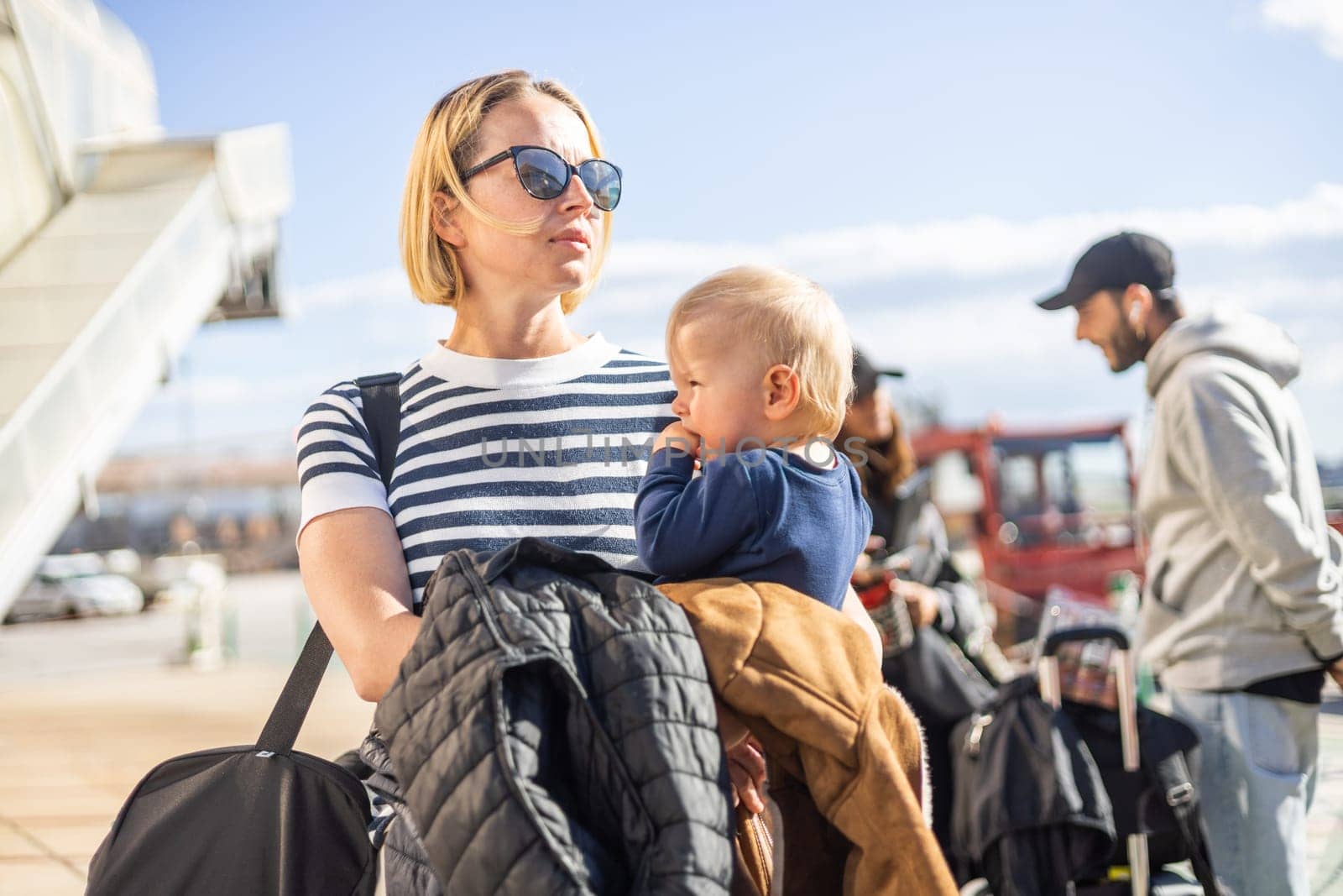 Motherat travelling with his infant baby boy child. Mom holding travel bag and her infant baby boy child while queuing for bus in front of airport terminal station