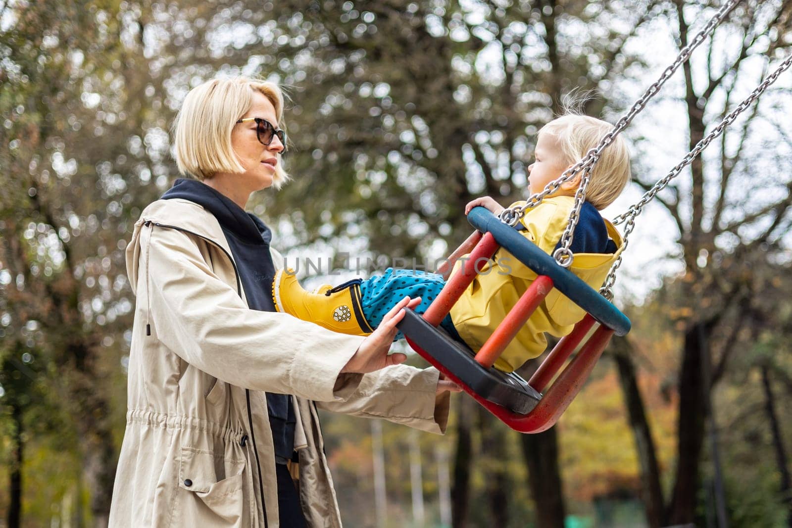 Mother pushing her infant baby boy child wearing yellow rain boots and cape on swing on playground outdoors on cold rainy overcast autumn day in Ljubljana, Slovenia.