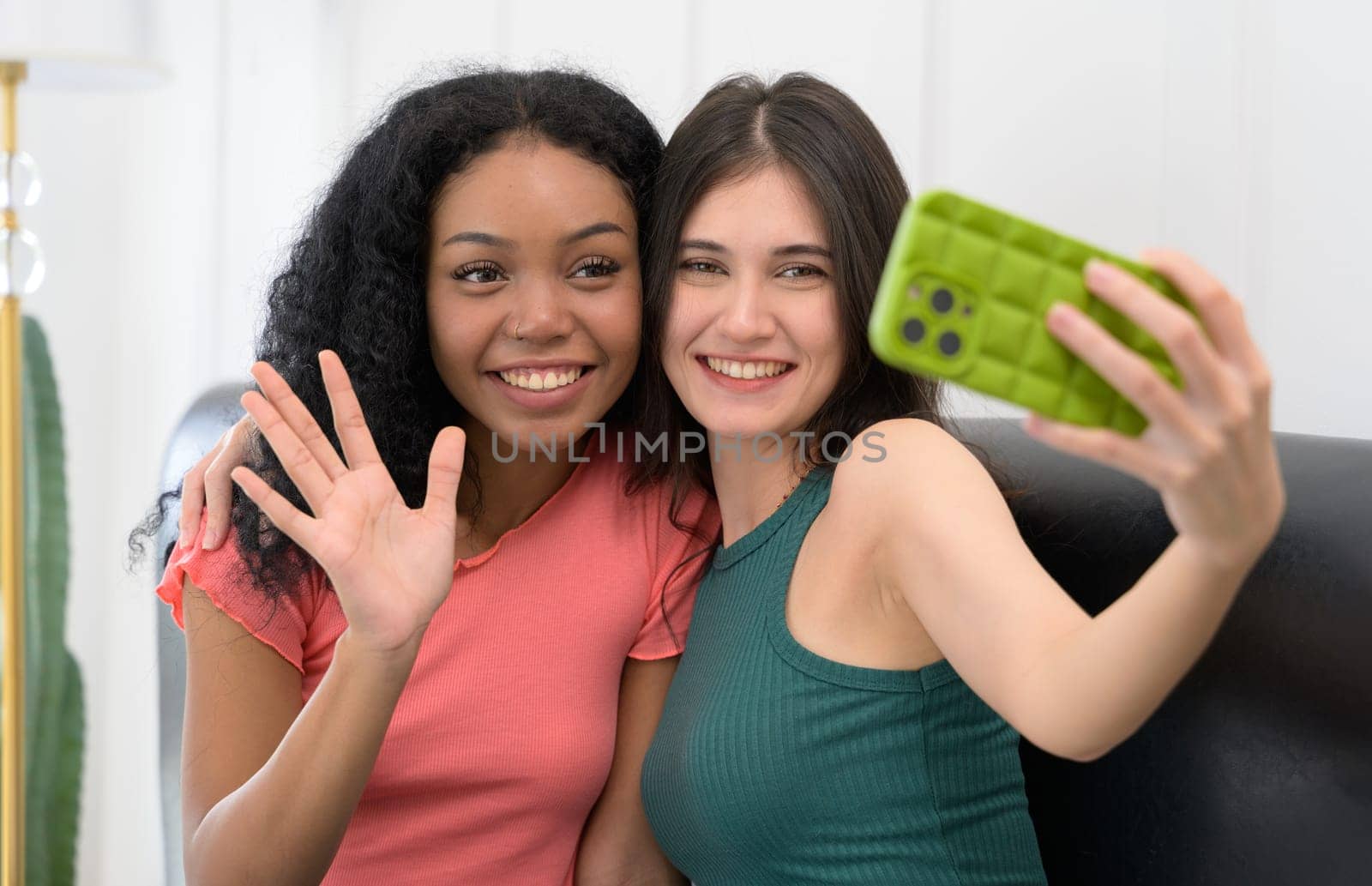 Two smiling young women taking a selfie with mobile phone at home. Caucasian girl in green t-shrit holding smartphone and African female raise a hand waving while sitting on couch.