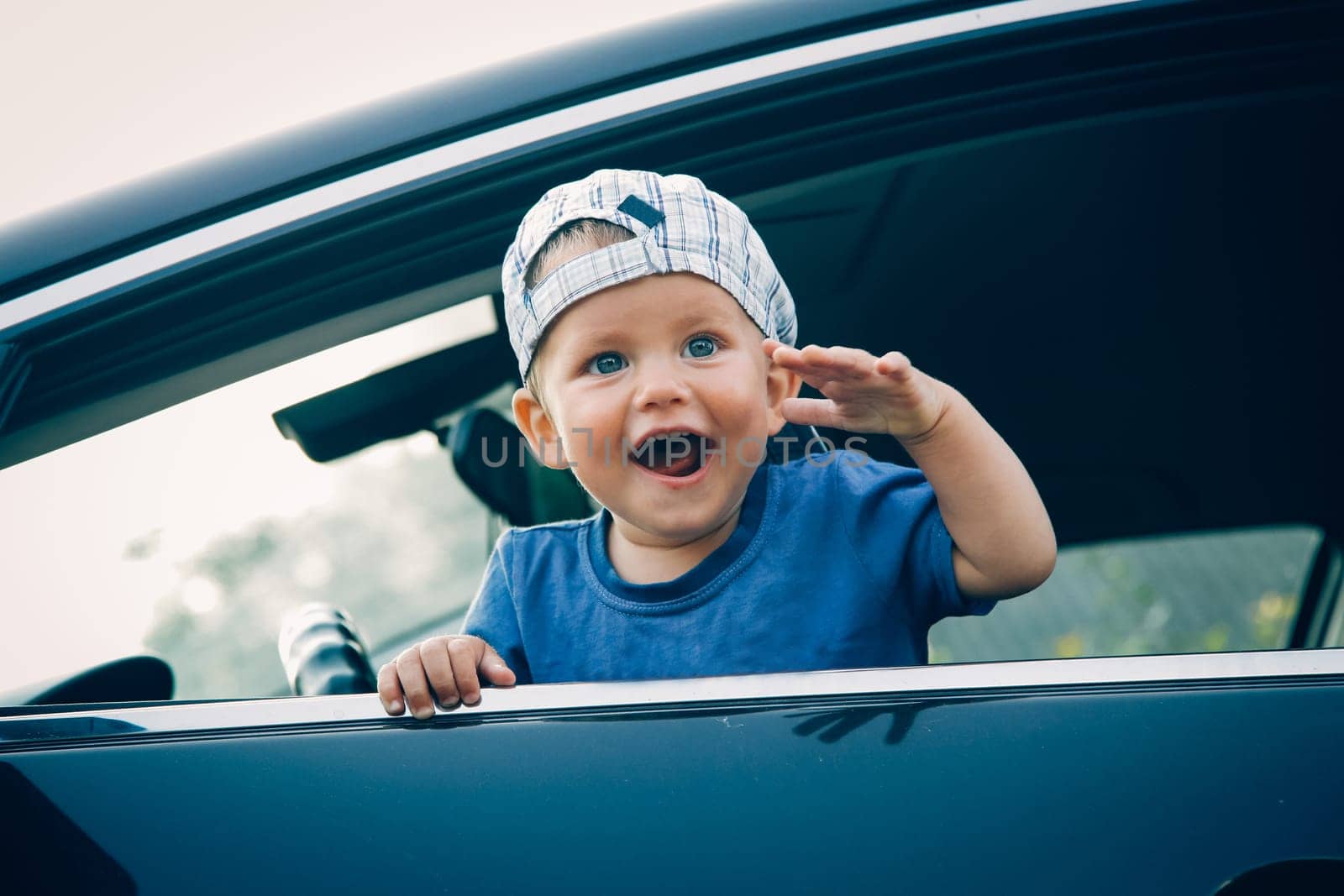 A small child looks out of the car window laughing happily and waving his hand