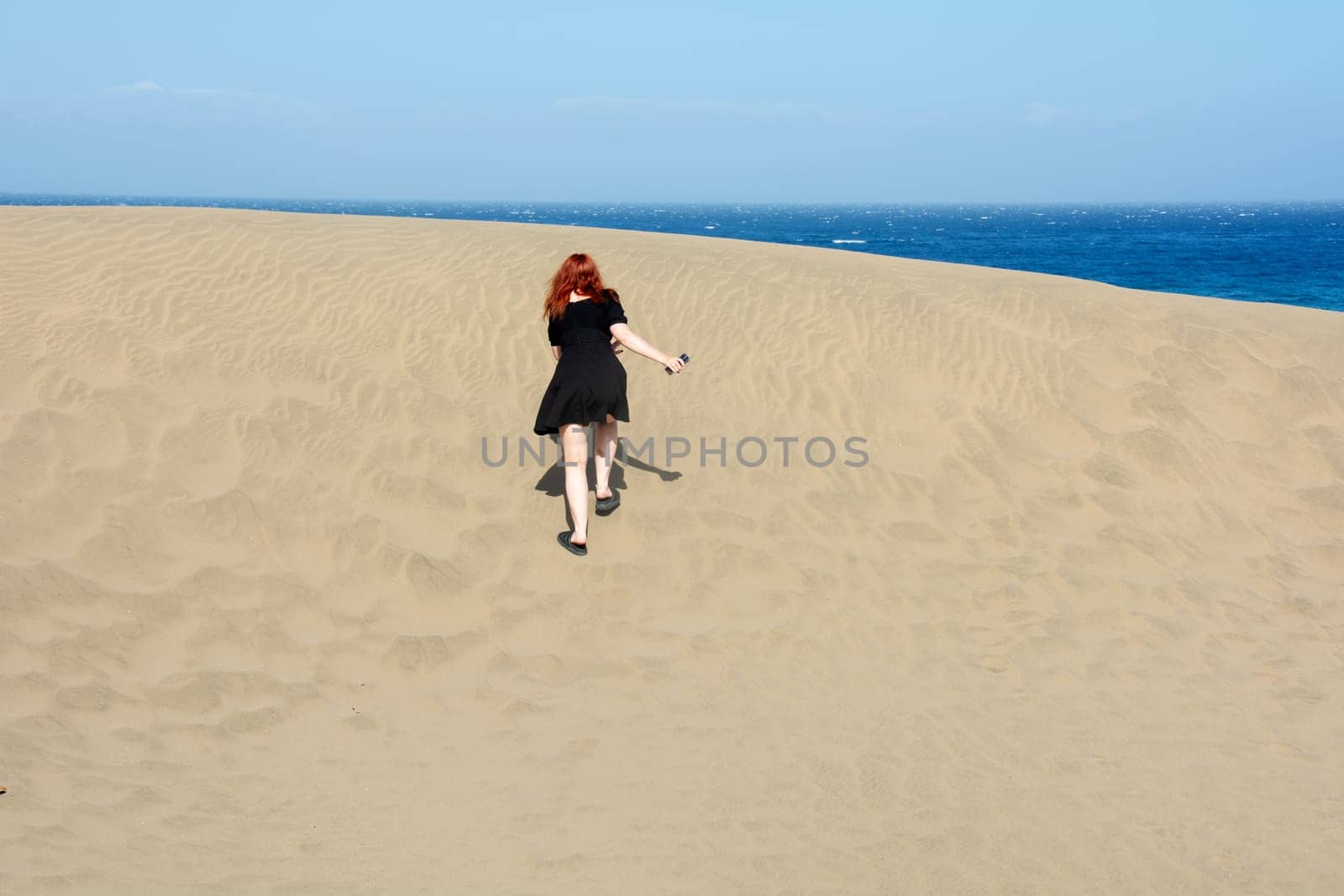 Young woman running up a sand dune towards the sea, with blue sky
