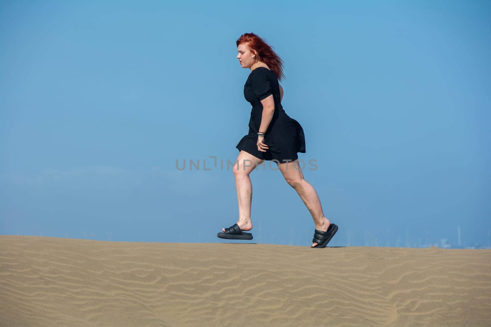 Young girl in a black short summer dress running on a sand dune with a blue sky