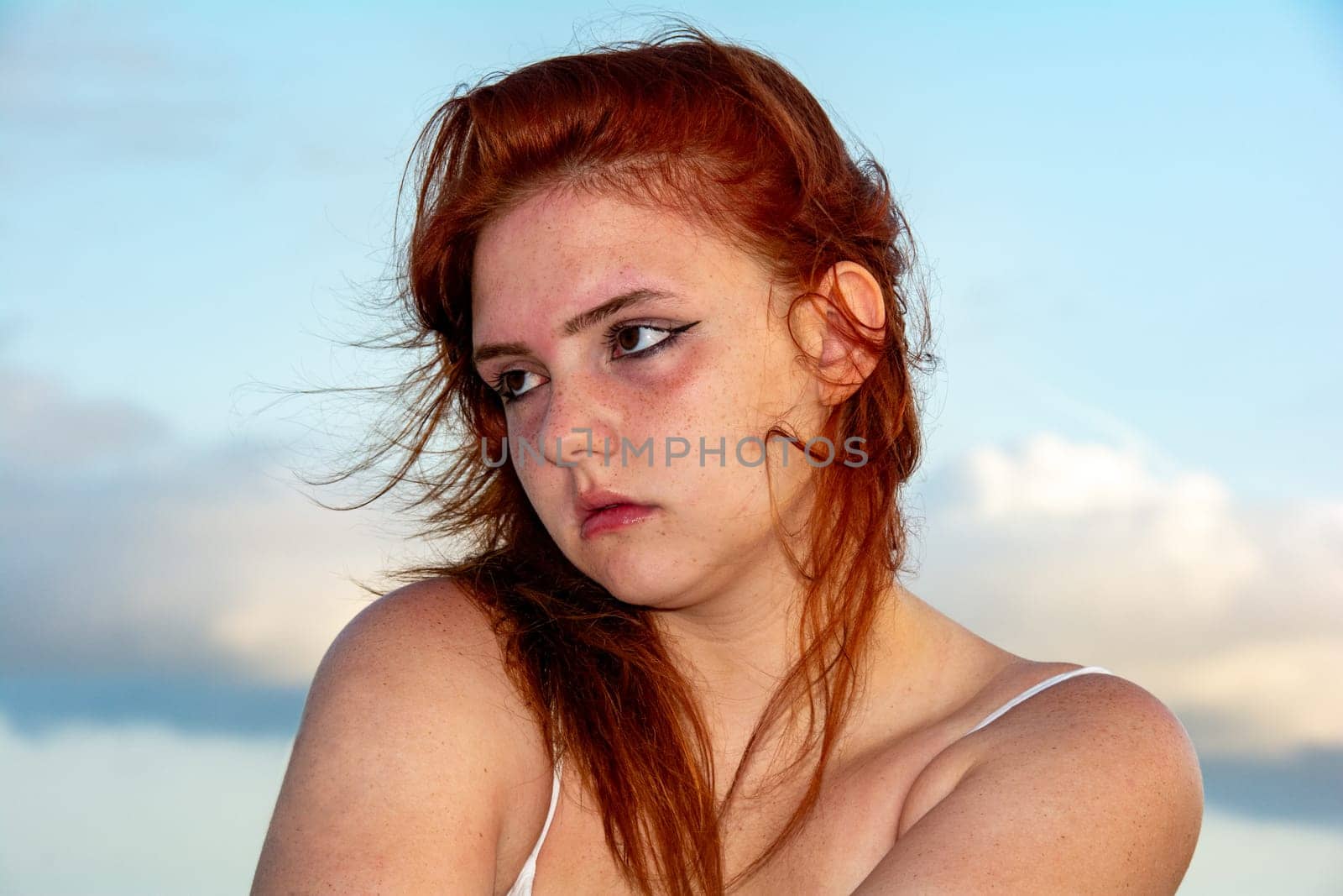 Portrait of a young girl with red hair, blue sky and clouds, hair tousled by the wind