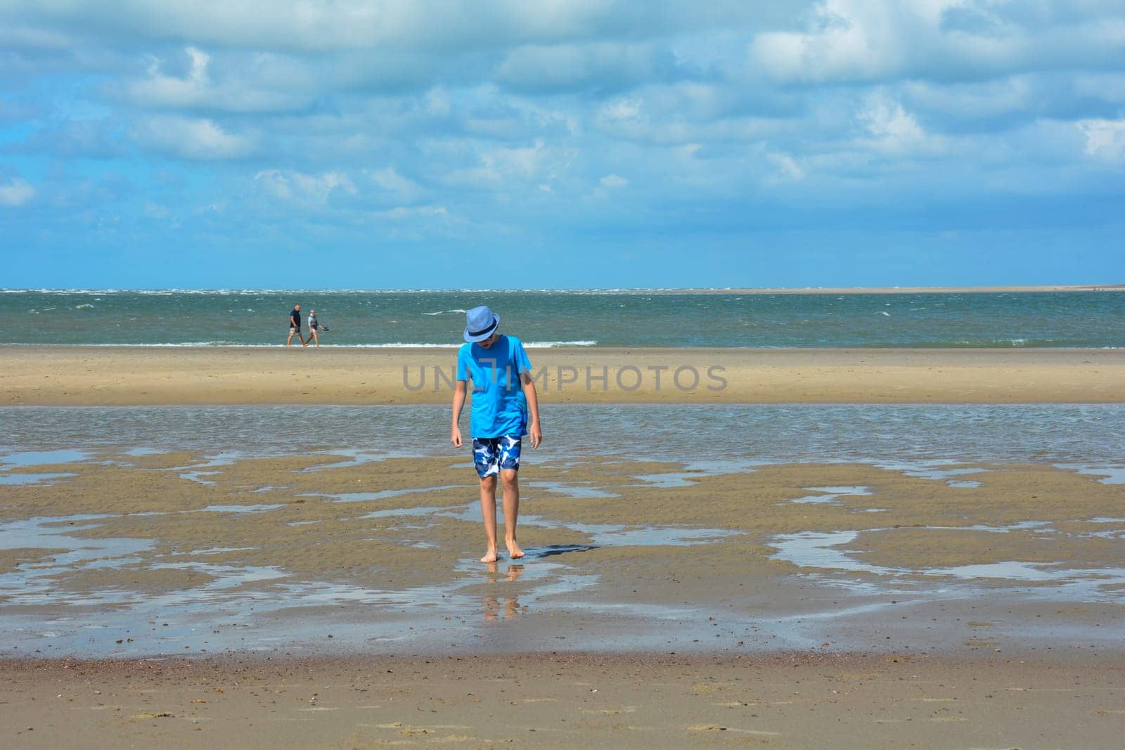 A boy in a hat runs on the sandy beach at low tide, with the sea and blue sky