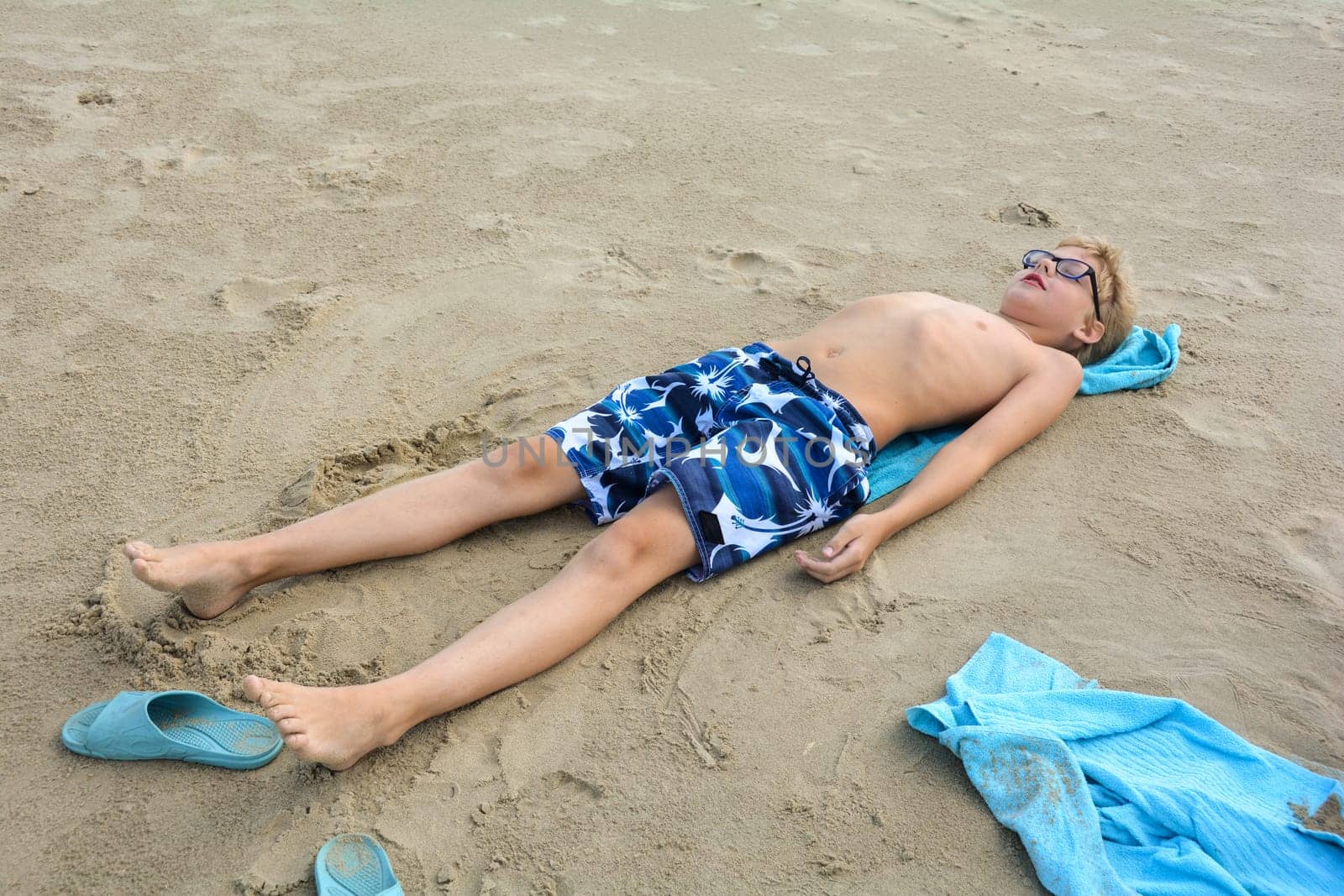 A boy with glasses lies on a towel on the beach to sunbathe in the sand