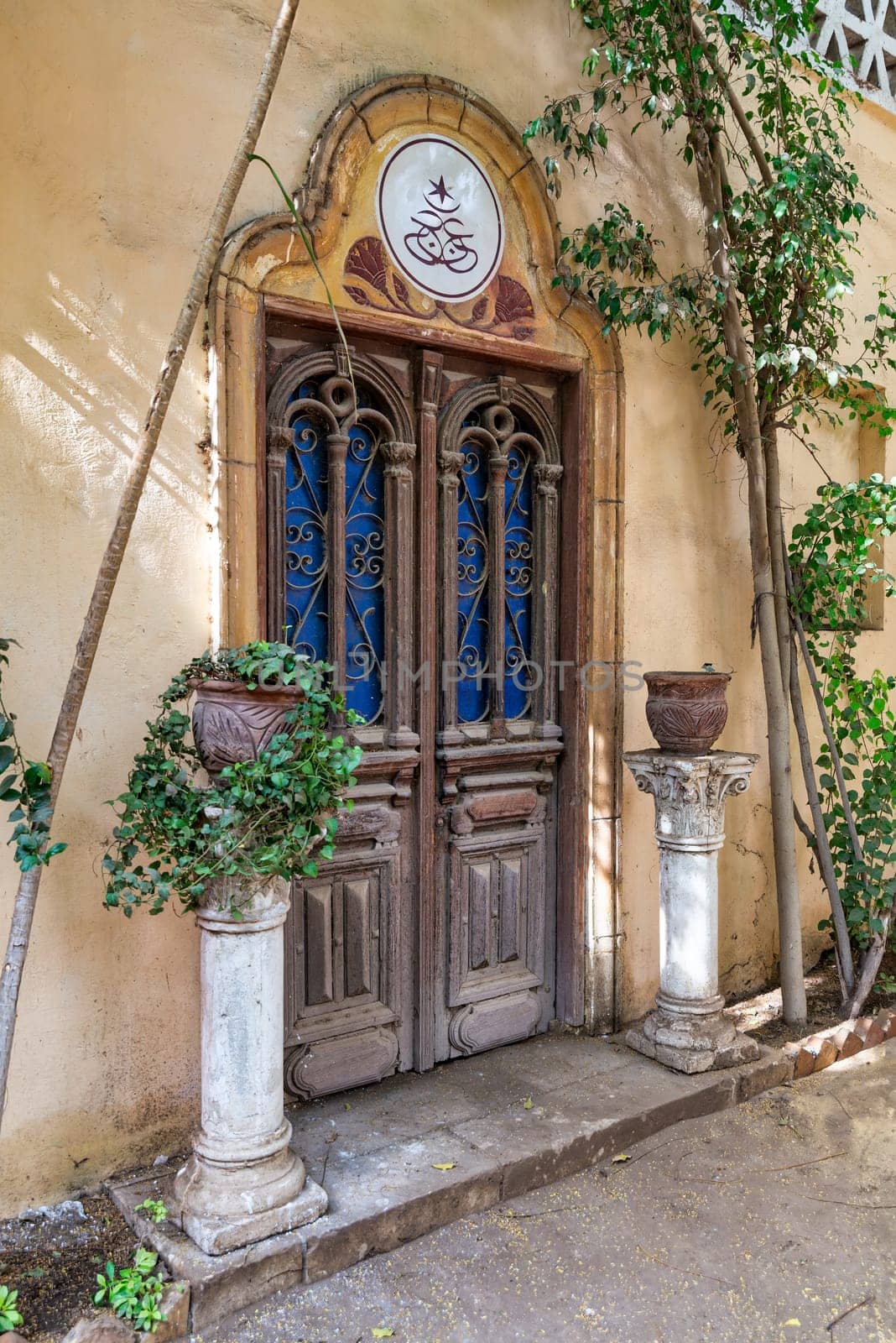 A closeup of Grungy traditional Egyptian style intricately carved wooden door, with wrought iron decorations in a yellow stone wall surrounded by bushes in flower pots