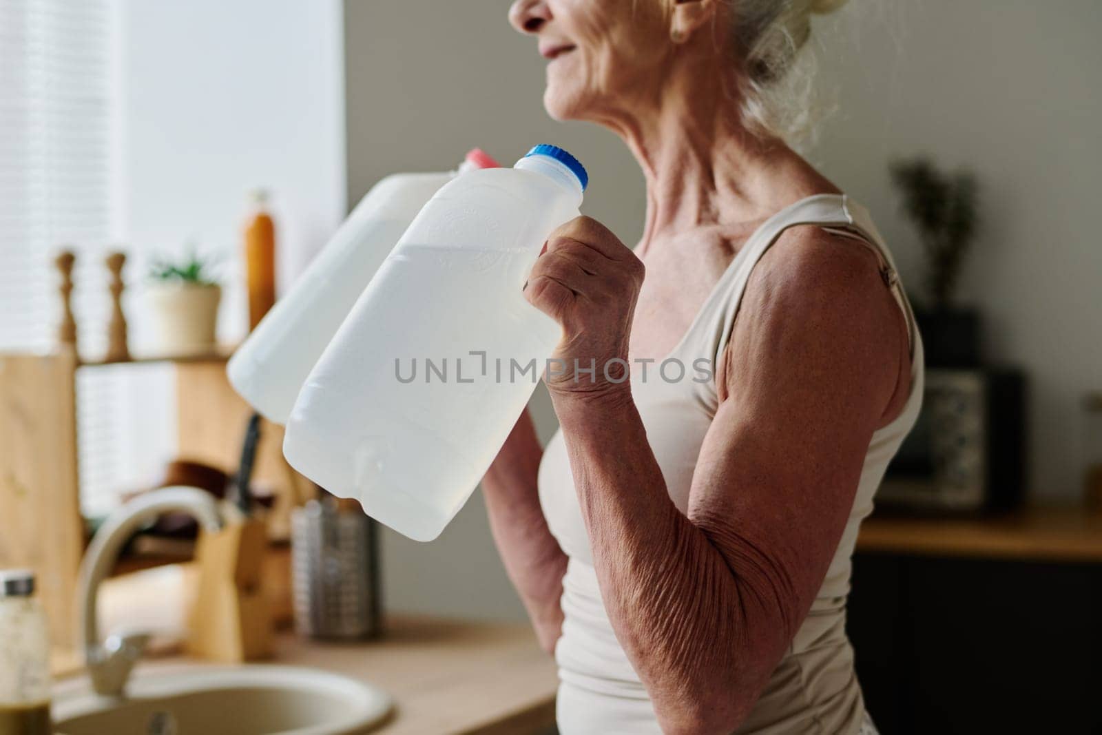 Cropped shot of senior woman in activewear doing physical exercise with two plastic canisters with water while standing in front of camera