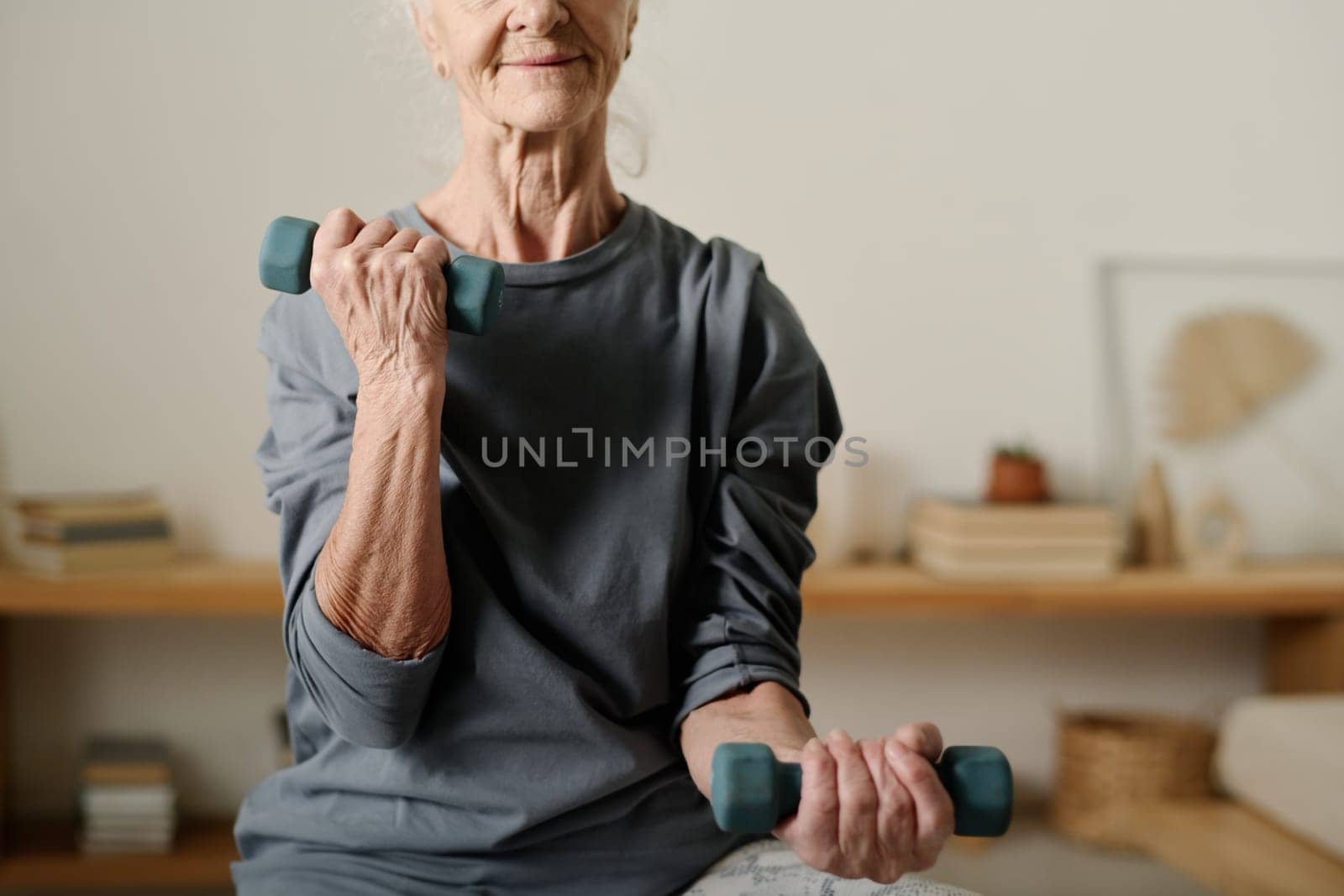 Active retired woman with dumbbells in hands pumping arm muscles during morning workout while sitting in front of camera
