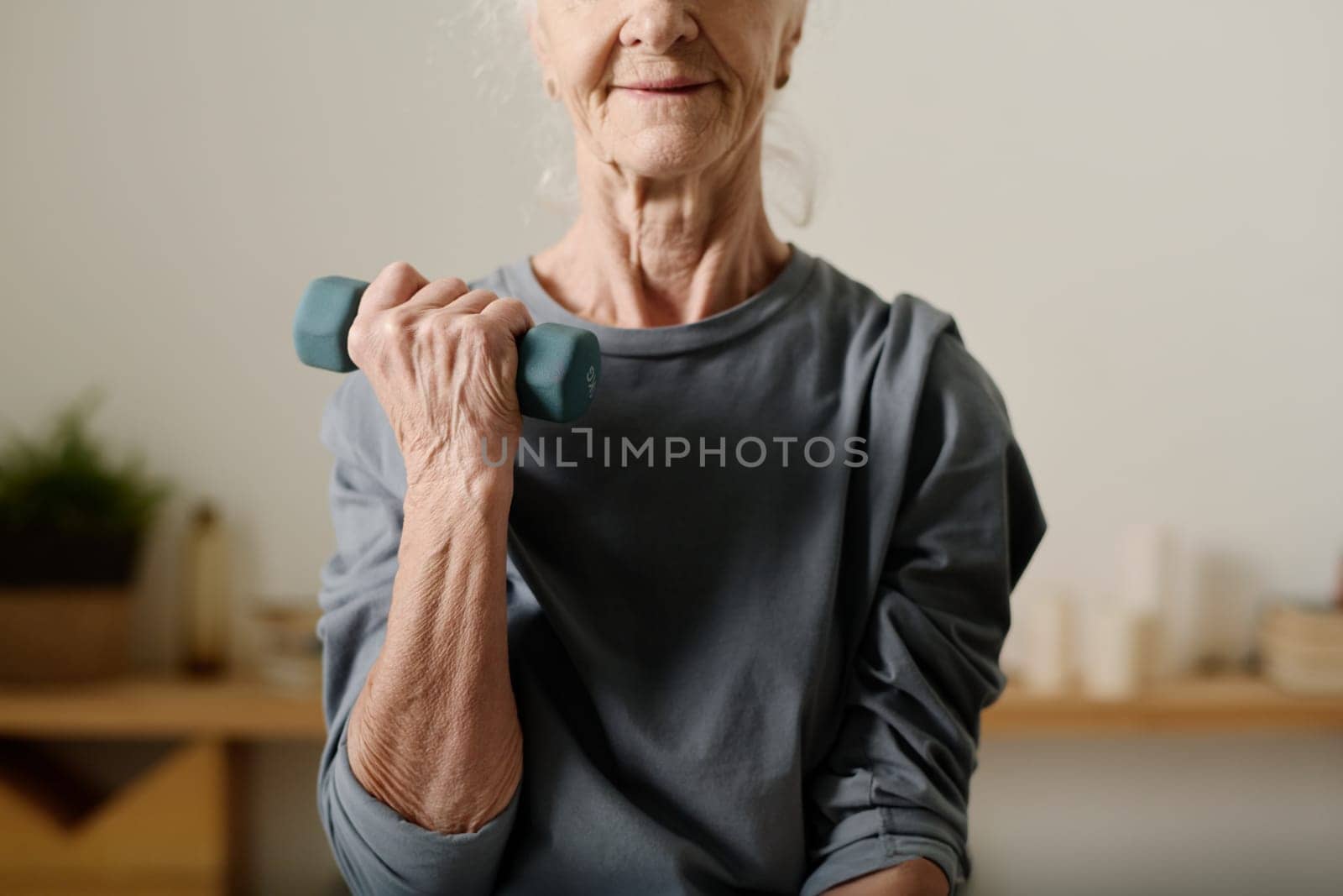 Cropped shot of senior active woman in grey cotton pullover sitting in front of camera at home and practicing exercise with dumbbells
