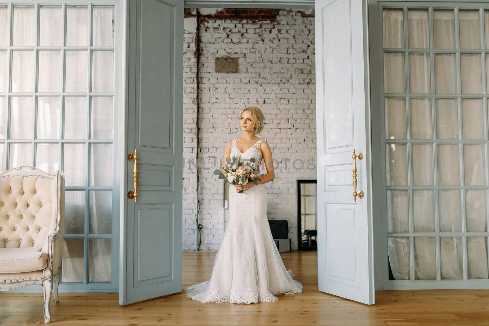 A blonde bride in a wedding dress and with a bouquet of flowers stands at the open door in the studio