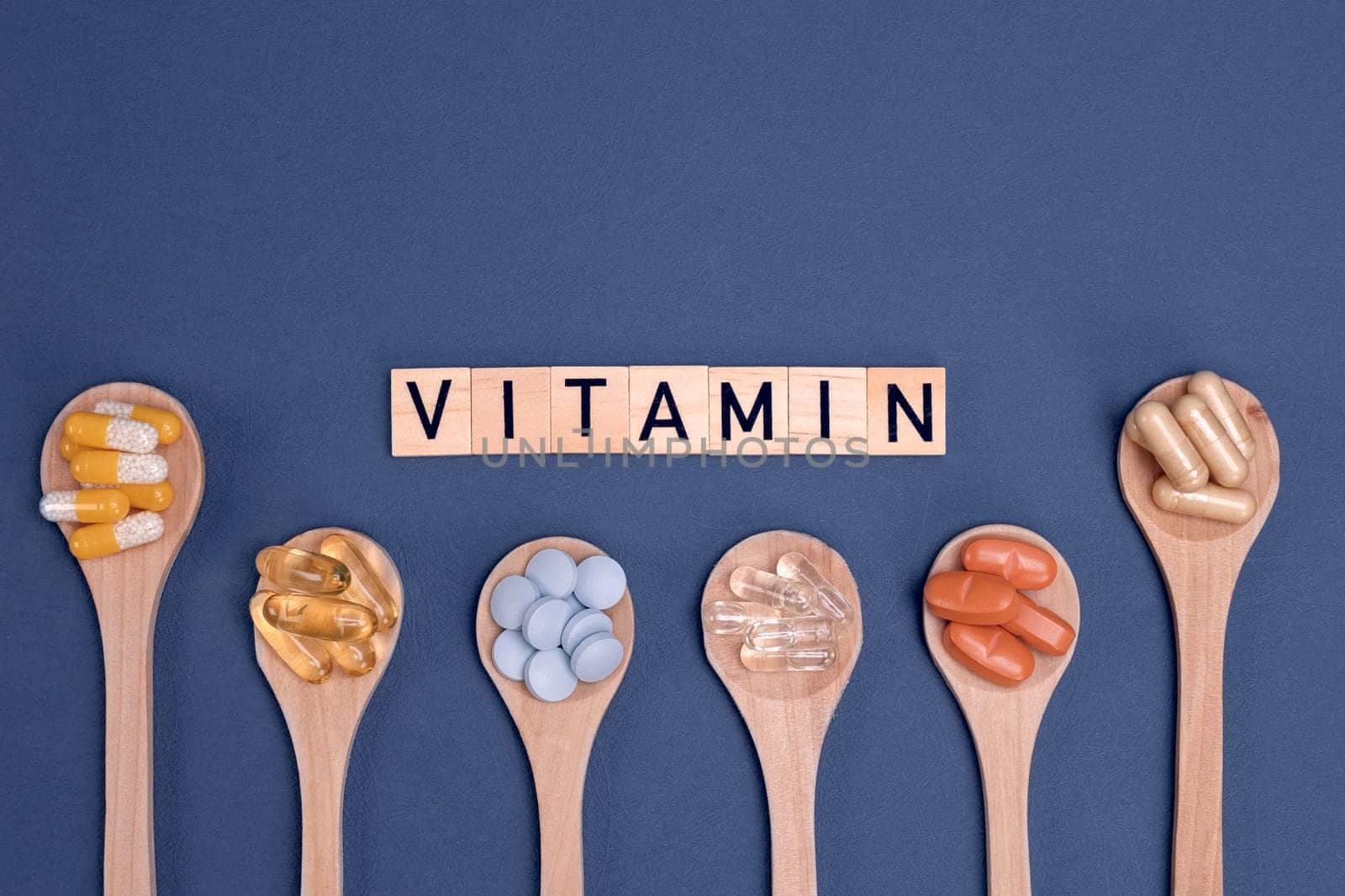 Different vitamins in wooden spoons with text on gray background. Pills, drugs, supplements, medicine, pharmacy and healthcare concept.