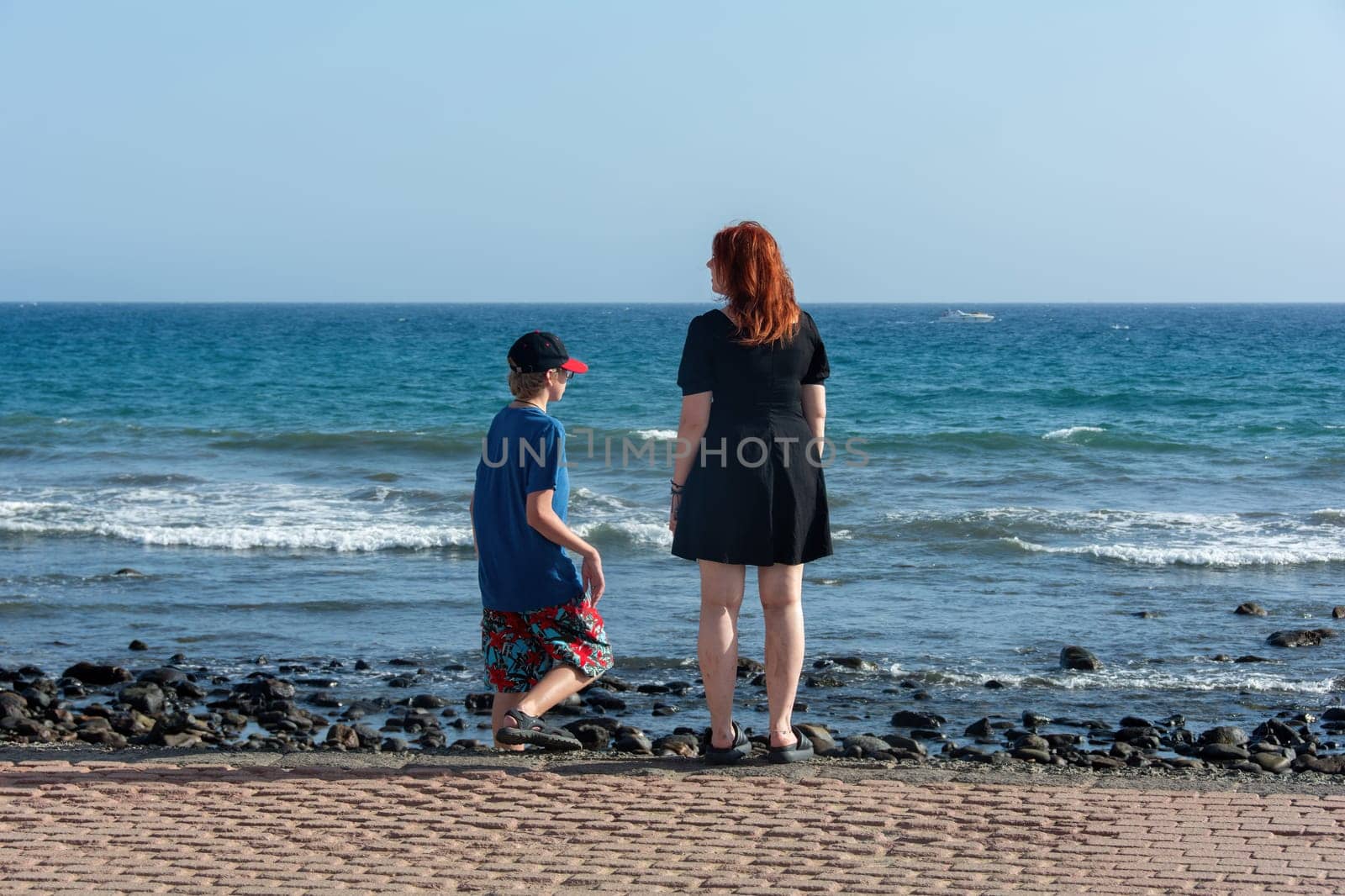 Young girl with a short black dress and a boy with shorts and a cap on a promenade by the sea, with blue sky,Young girl with a short black dress and a boy with shorts and a cap on a promenade by the sea, with blue sky