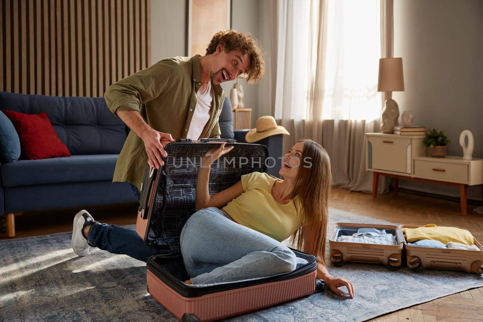 Overjoyed playful family couple preparing for vacation fooling around while packing clothing stuff. Young man putting woman into suitcase. Happy people celebrating travel start