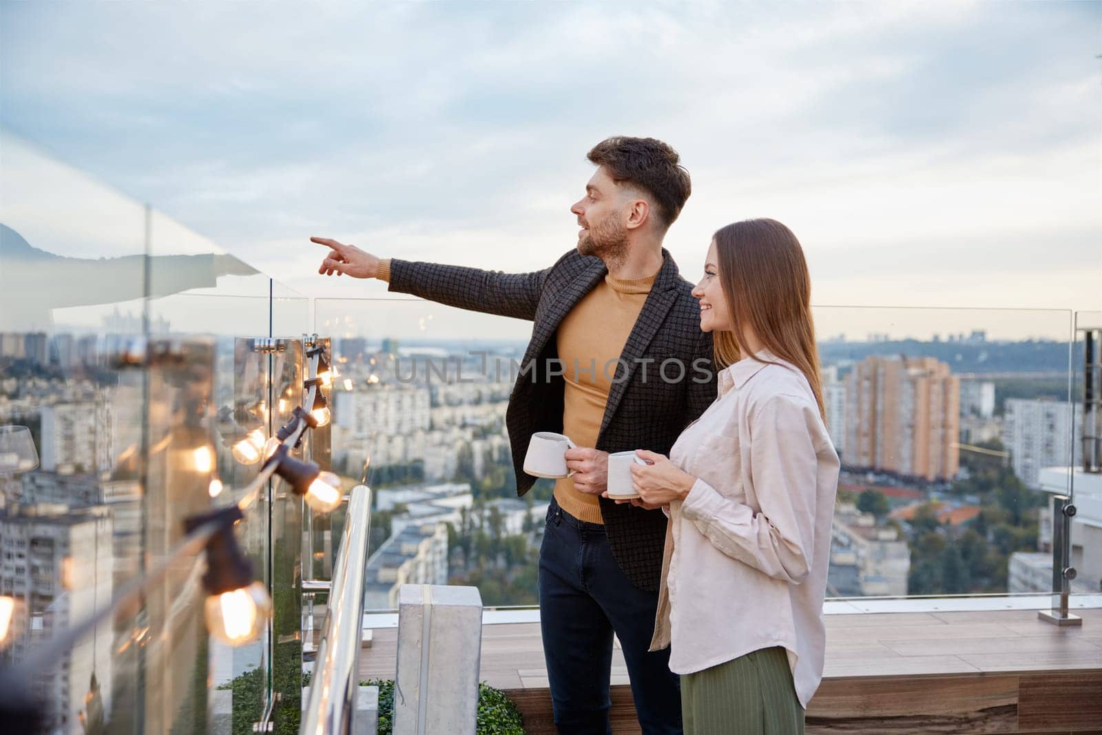 Business colleagues chitchatting during coffee break at office roof terrace. Young man project manager pointing to city building discussing real estate