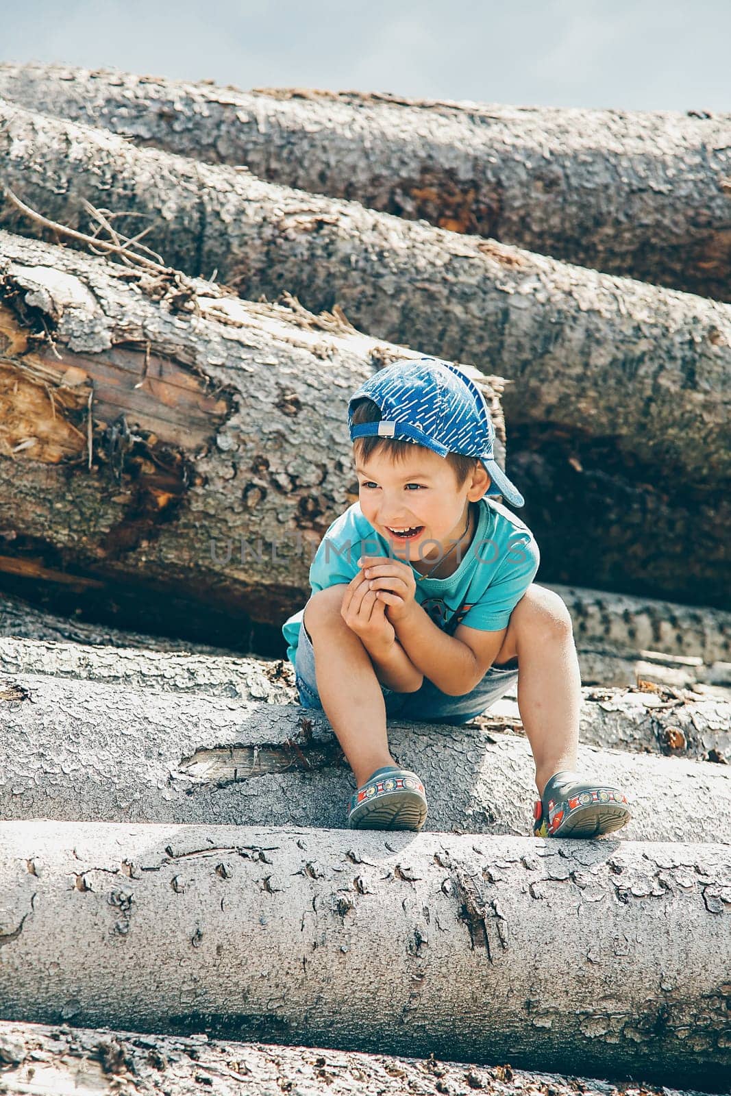 A little boy in a blue baseball cap and short shorts sits on a log during the day and laughs merrily