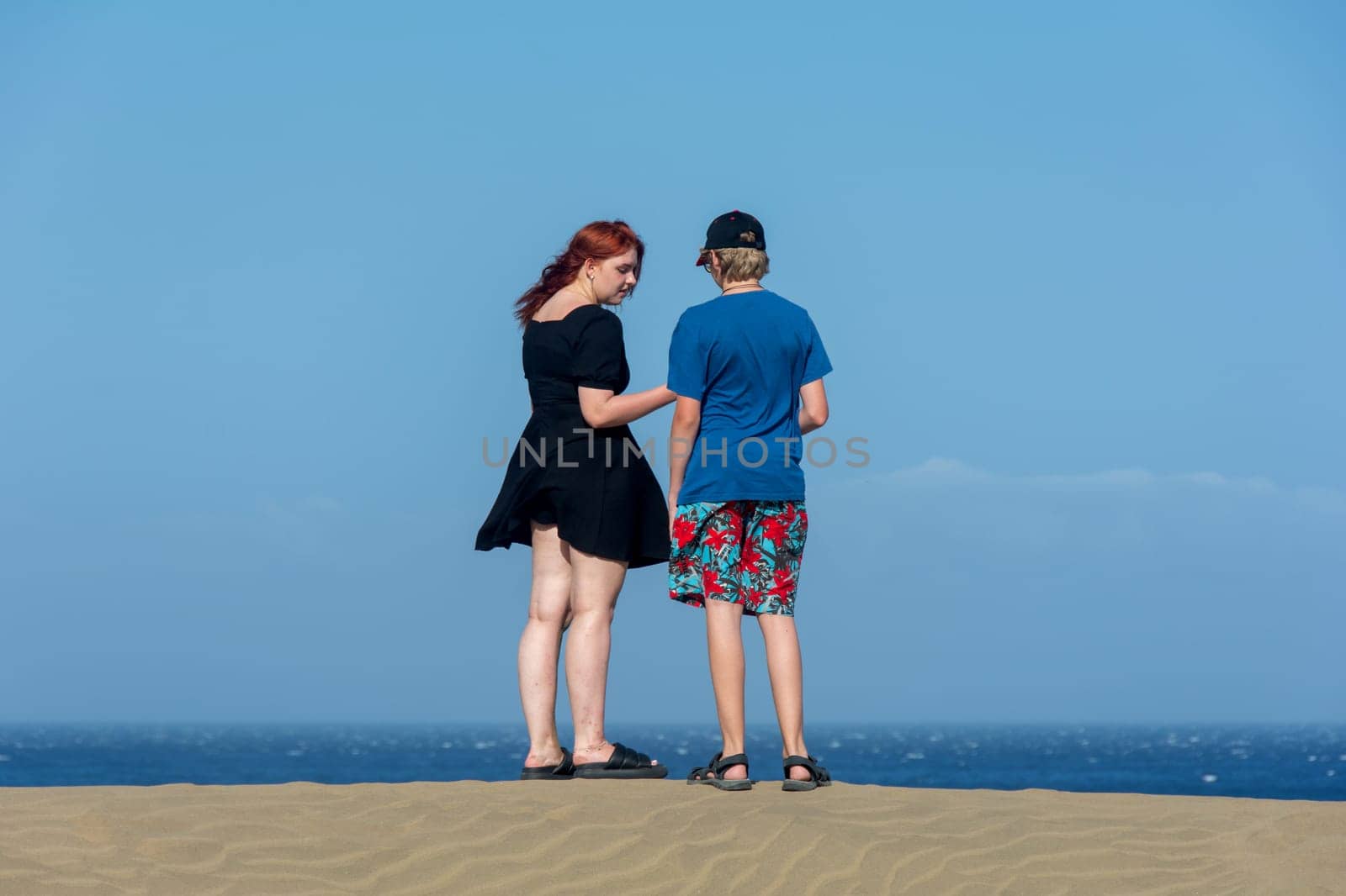Two teenagers stand on a sand dune, with sea and blue sky