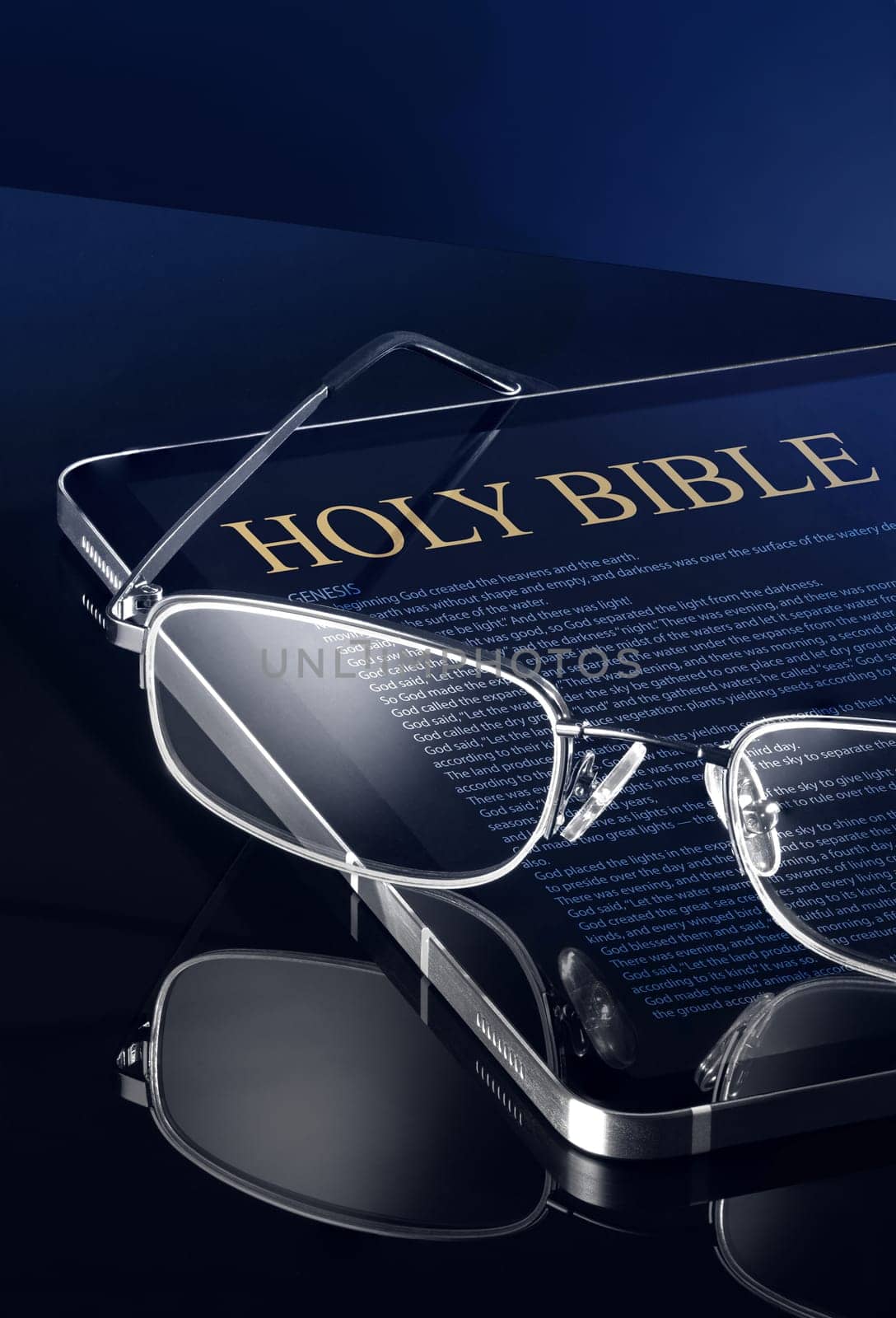 This close-up image shows a tablet, either Android or iOS, with the words Holy Bible and a passage from the Bible written on it. A pair of reading glasses is placed on top of the tablet. The tablet and glasses are on a shiny dark blue table, and they are not entirely in the frame.