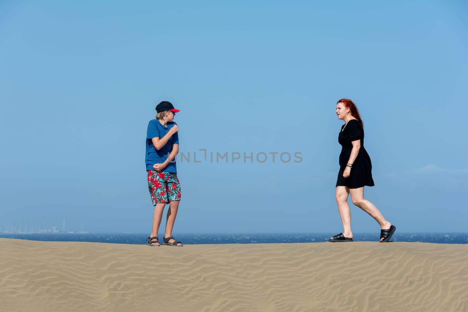 Two teenagers stand on a sand dune, with sea and blue sky