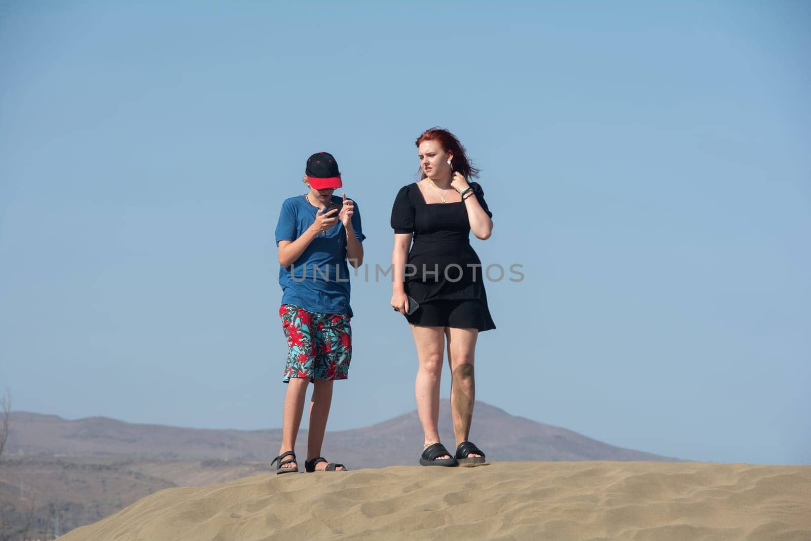 Two teenagers are standing on a sand dune with their cell phones, the boy is looking at his smartphone