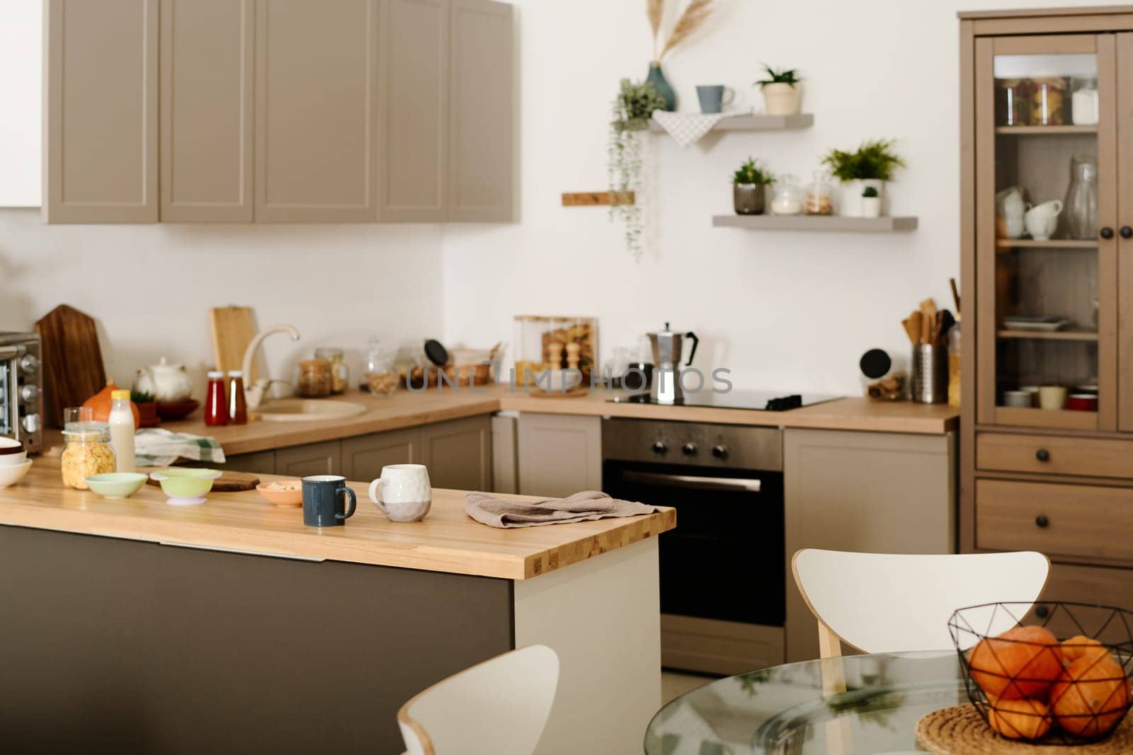 Part of modern spacious kitchen with row of cabinets hanging over counter with kitchenware and sink and electric stove with coffee pot
